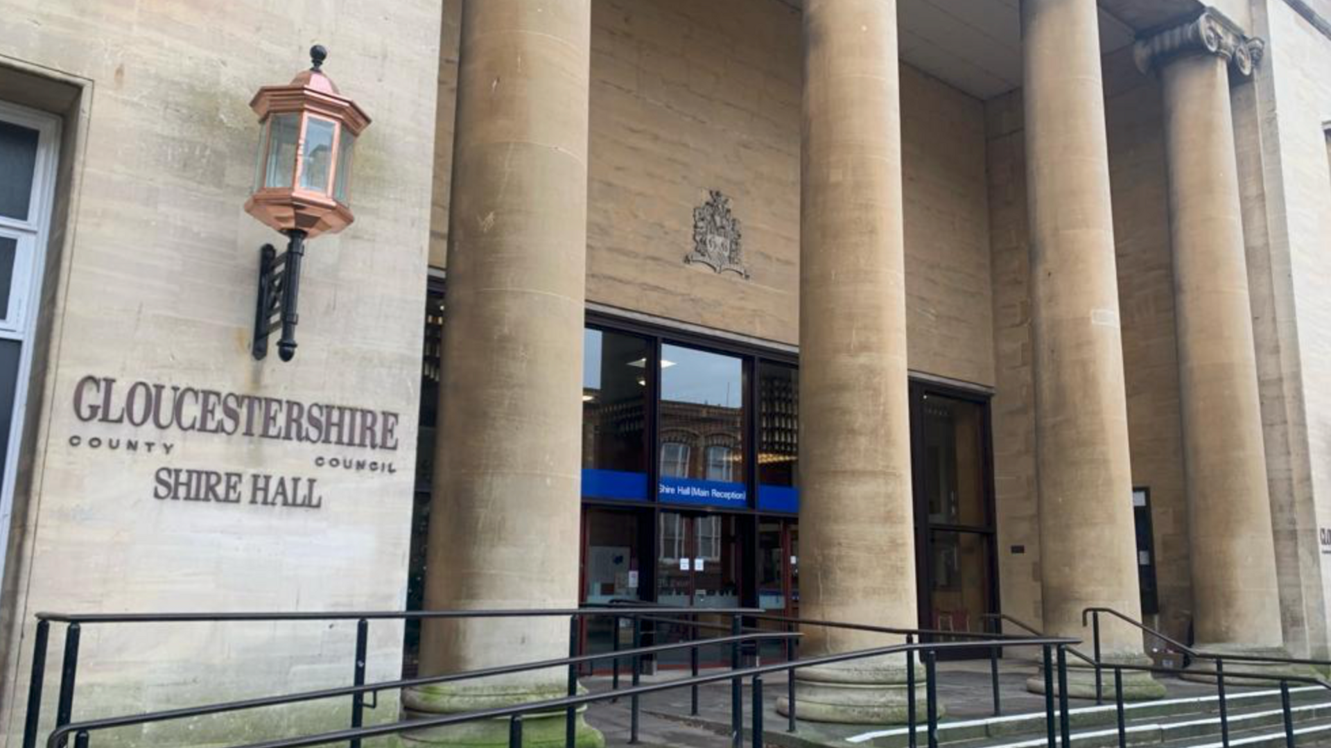 Shire Hall in Gloucester. There are four large stone pillars at the glass door entrance to the building. On the left is tiling reading 'Gloucestershire County Council Shire Hall", with a copper street lamp above it.