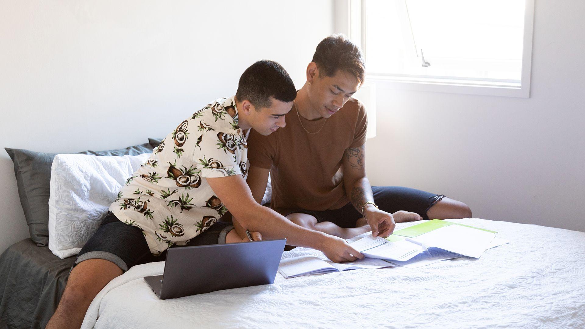 Stock photo shows a couple stiiting on a bed in their home looking at a laptop and bills.