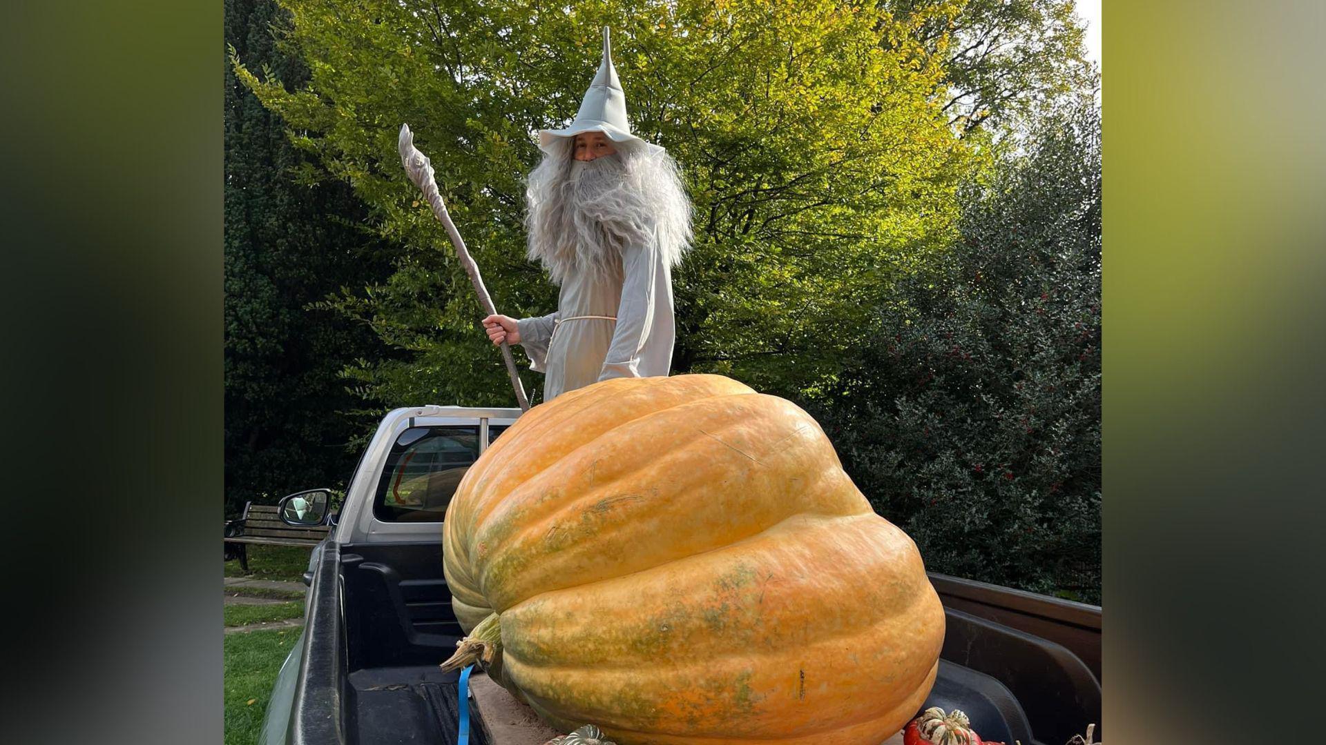 A man dressed in a grey wizard's costume. He is standing at the back of a utility vehicle, with a giant pumpkin and holding a staff. 