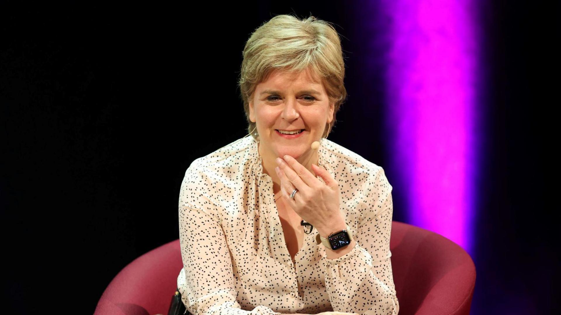 Nicola Sturgeon sitting in a red chair and laughing. She is wearing a white blouse with black dots on it and a jar of water is on the table in front of her