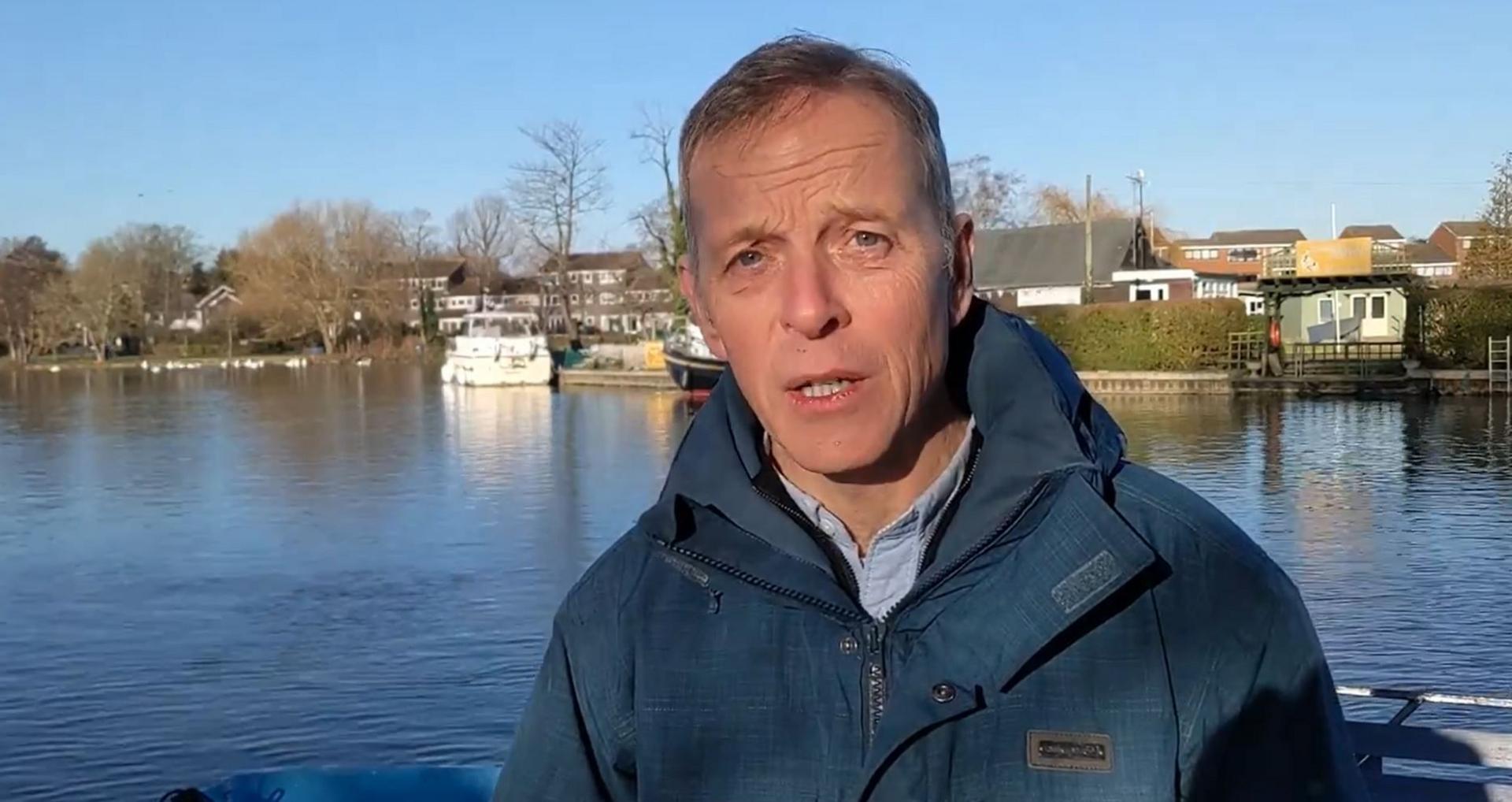 Matt Rodda stands in front of a lake looking at the camera. He has a concerned look on his face and looks as though he is mid-speech. Mr Rodda has short greying hair and wears a pale blue collared shirt under a deeper blue waterproof collared jacket. Behind him is a lake, lined by a row of houses, trees and hedging.