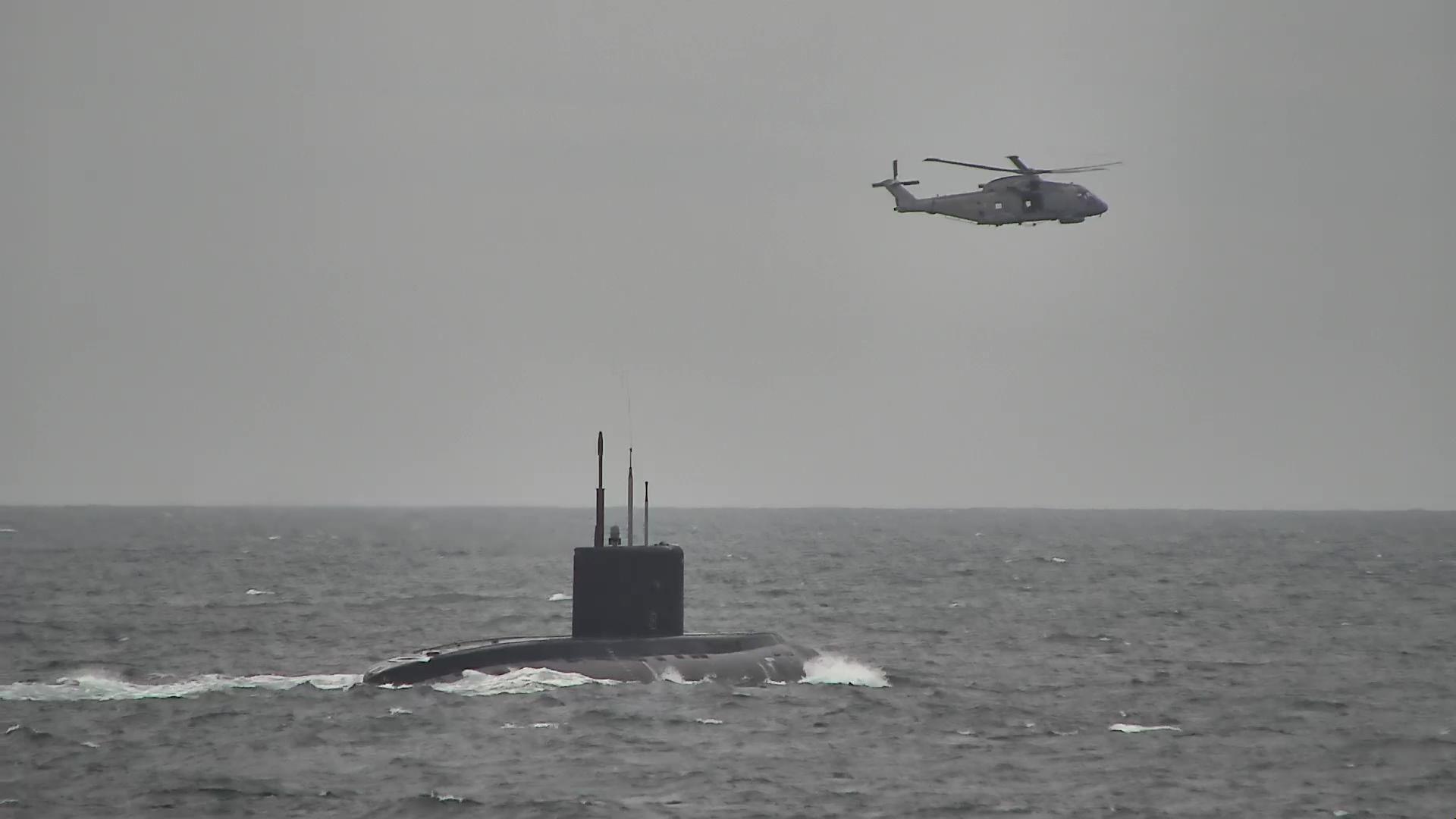 A submarine at the front of the image in the water with a helicopter flying in the sky above
