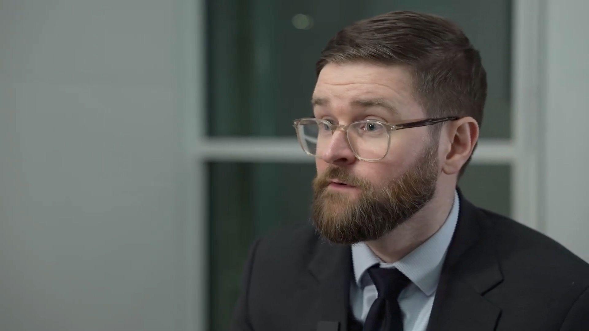 David Palmer speaks off camera, he has a beard and glasses and is wearing a suit