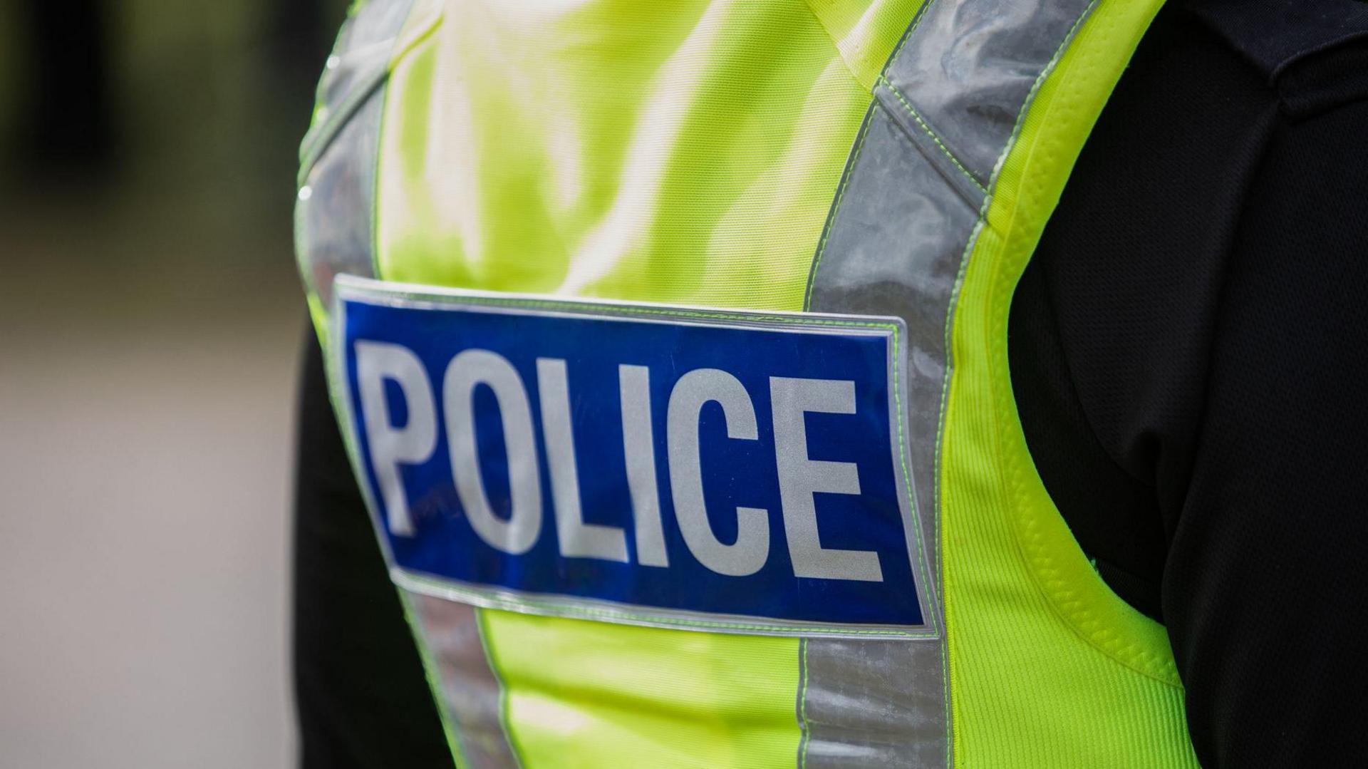 The back of a police officer, who is wearing a hi-vis jacket which has "police" written on the back of it