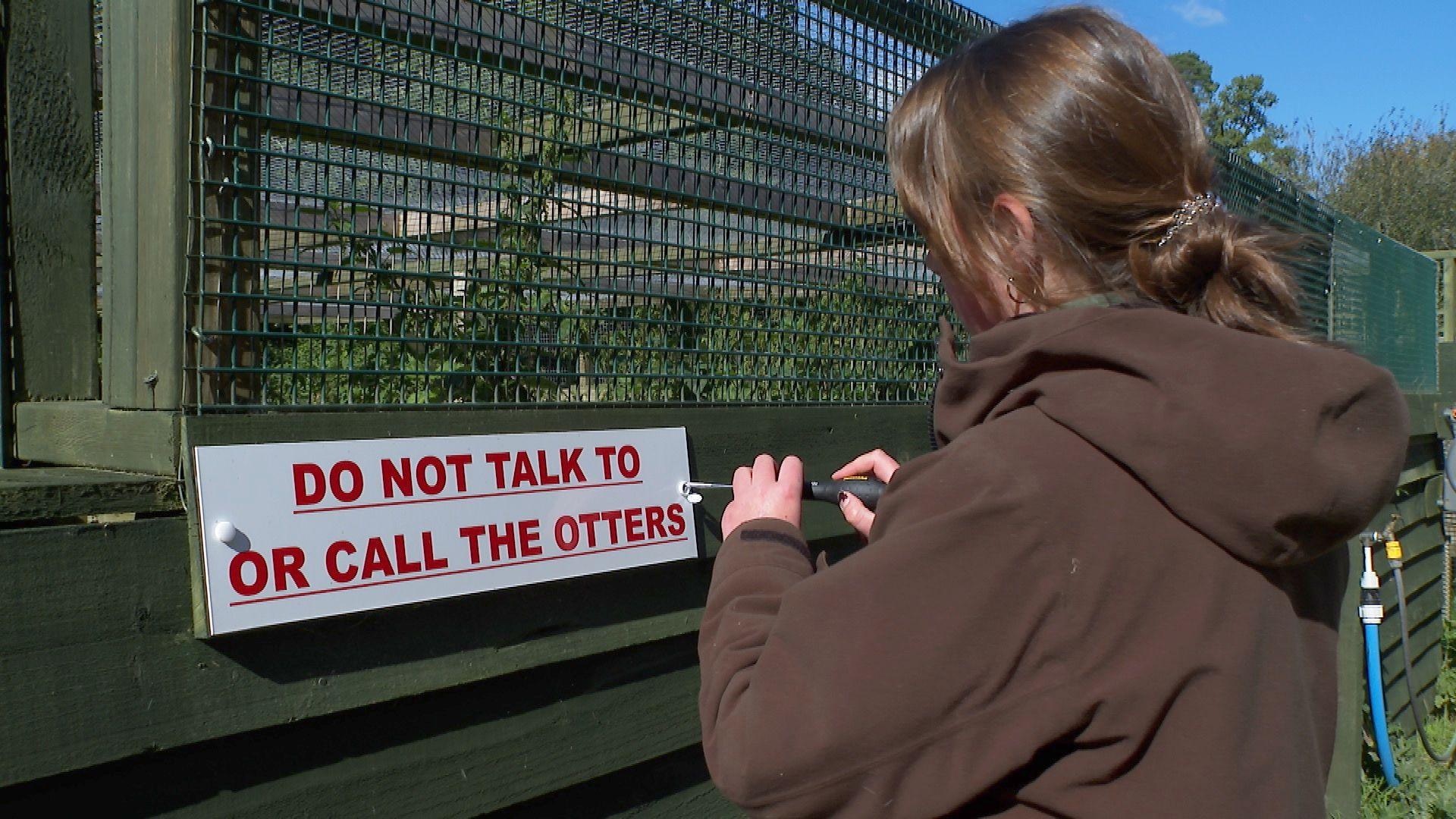 A woman dressed in a brown hoodie with her hair tied back is unscrewing a sign saying "Do not talk to or call the otters"