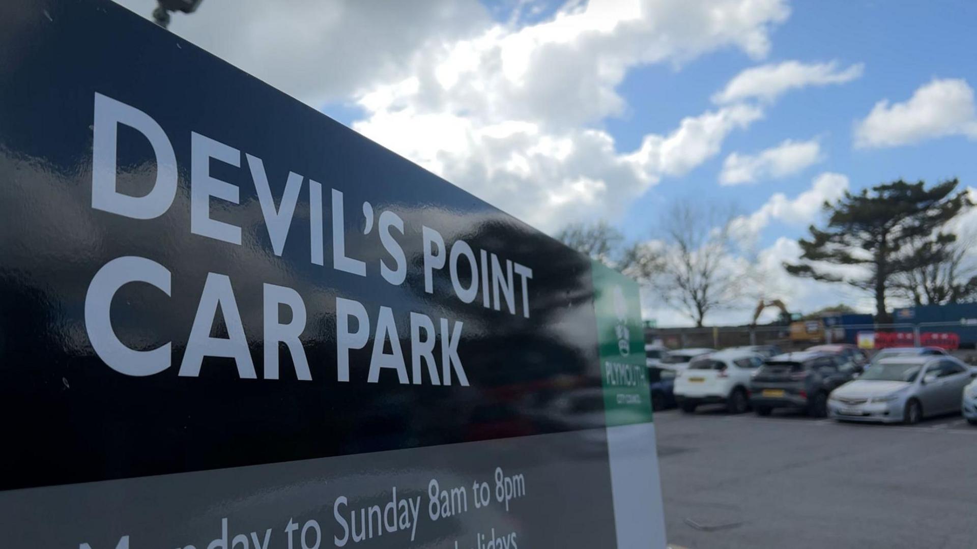 A close up picture of a car park sign with some cars in the background. The words 'Devil's Point Car Park' is visible with the green and white council logo also on it.