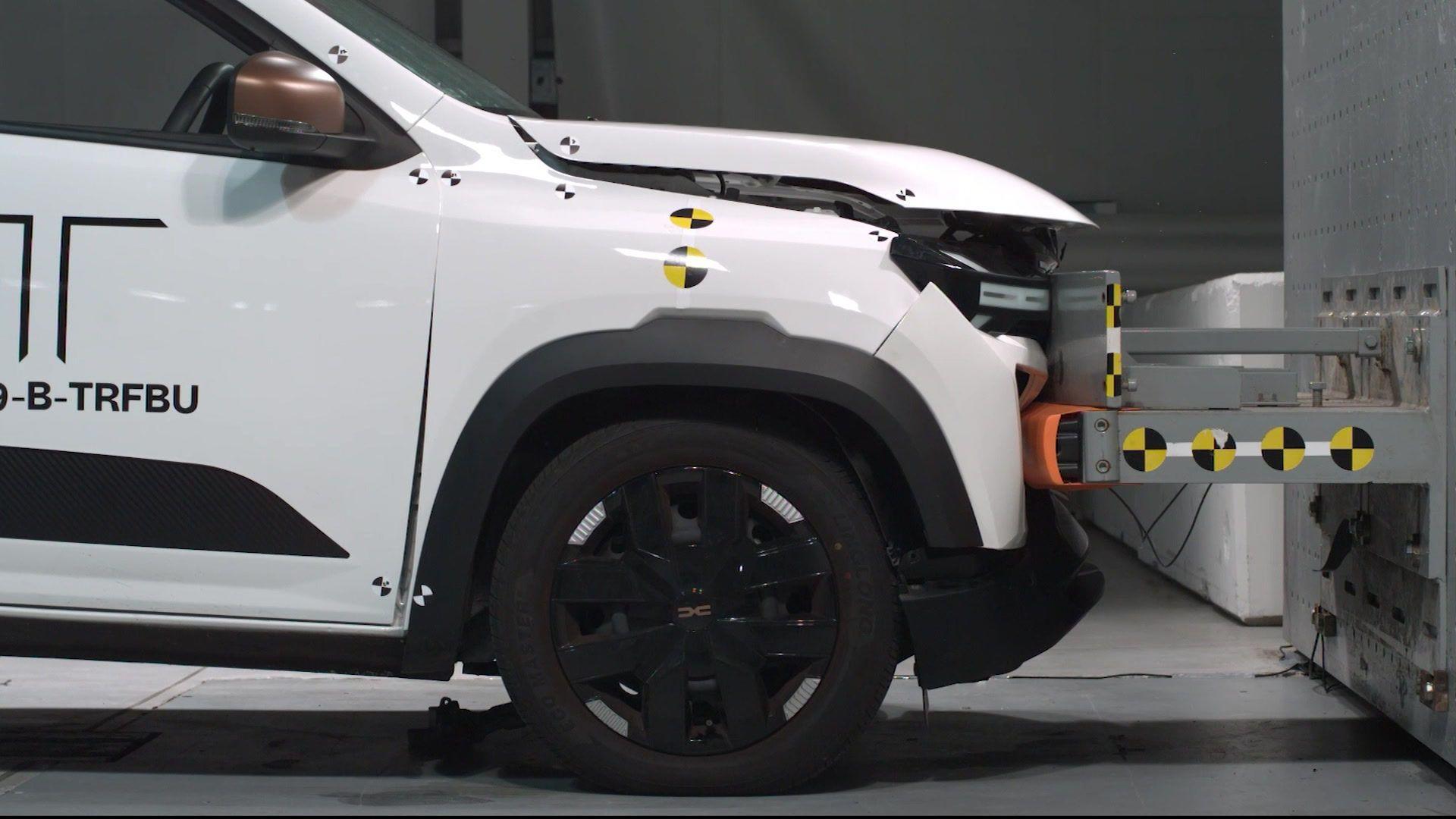 A white electric car crashing into a wall at a low speed in a crash test lab. The front of the car has bent in a bit and the bonnet has lifted up, but otherwise the damage looks minimal.