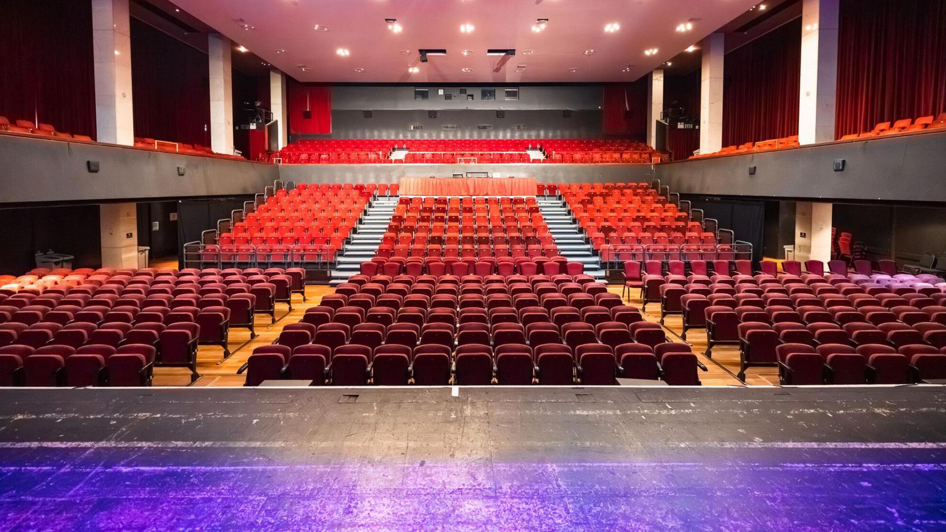 An empty theatre auditorium viewed from the stage, showing rows of red seats