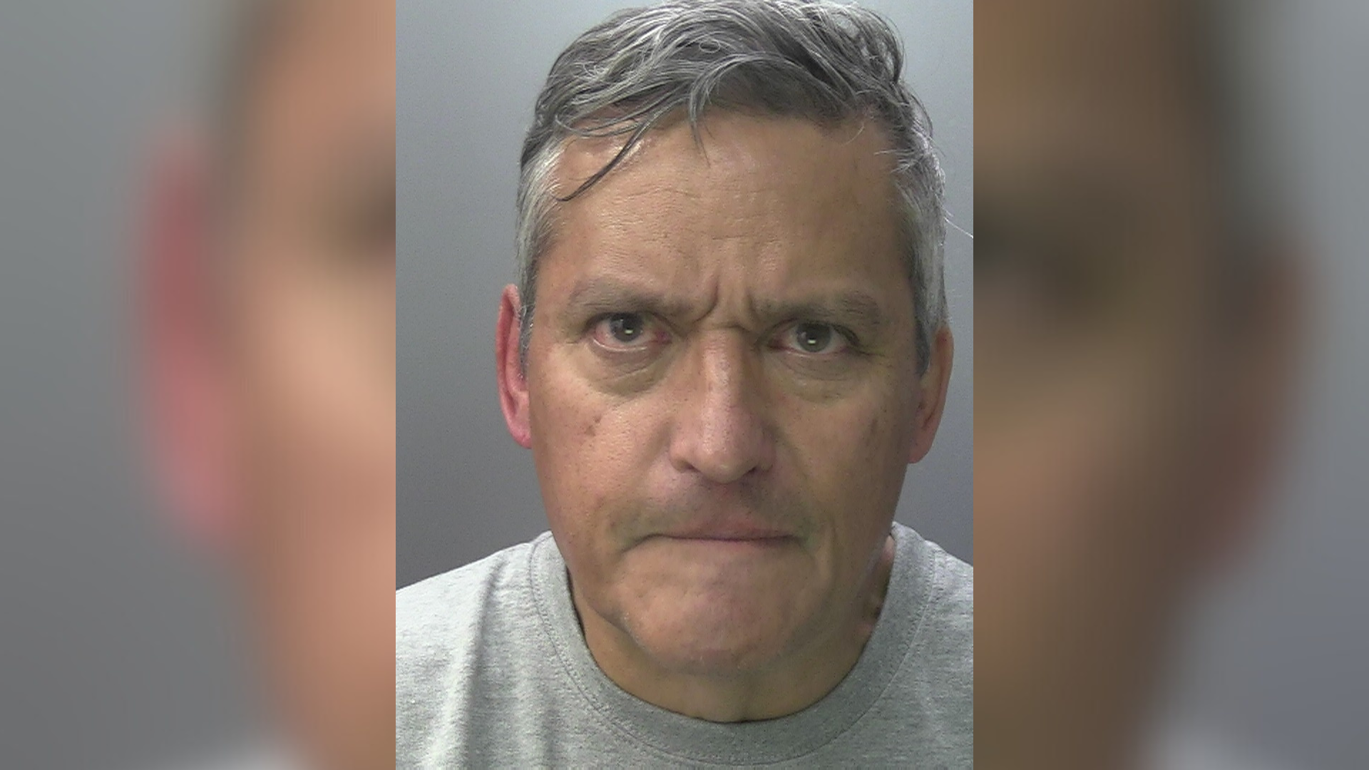 A man with short grey hair and a stern expression wearing a grey t-shirt in a police custody picture.