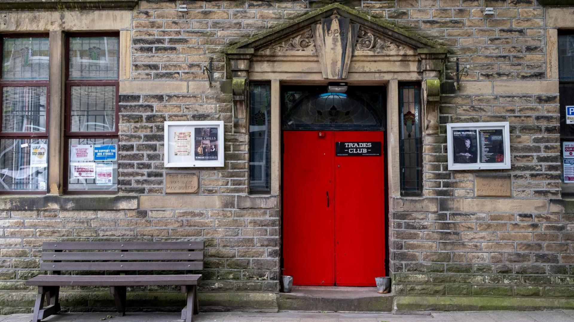 A red door at the front of a brick-built building with a small, black sign saying Trades Club on it in white writing. They are a number of posters for events pinned to notice boards on either side of the doorway