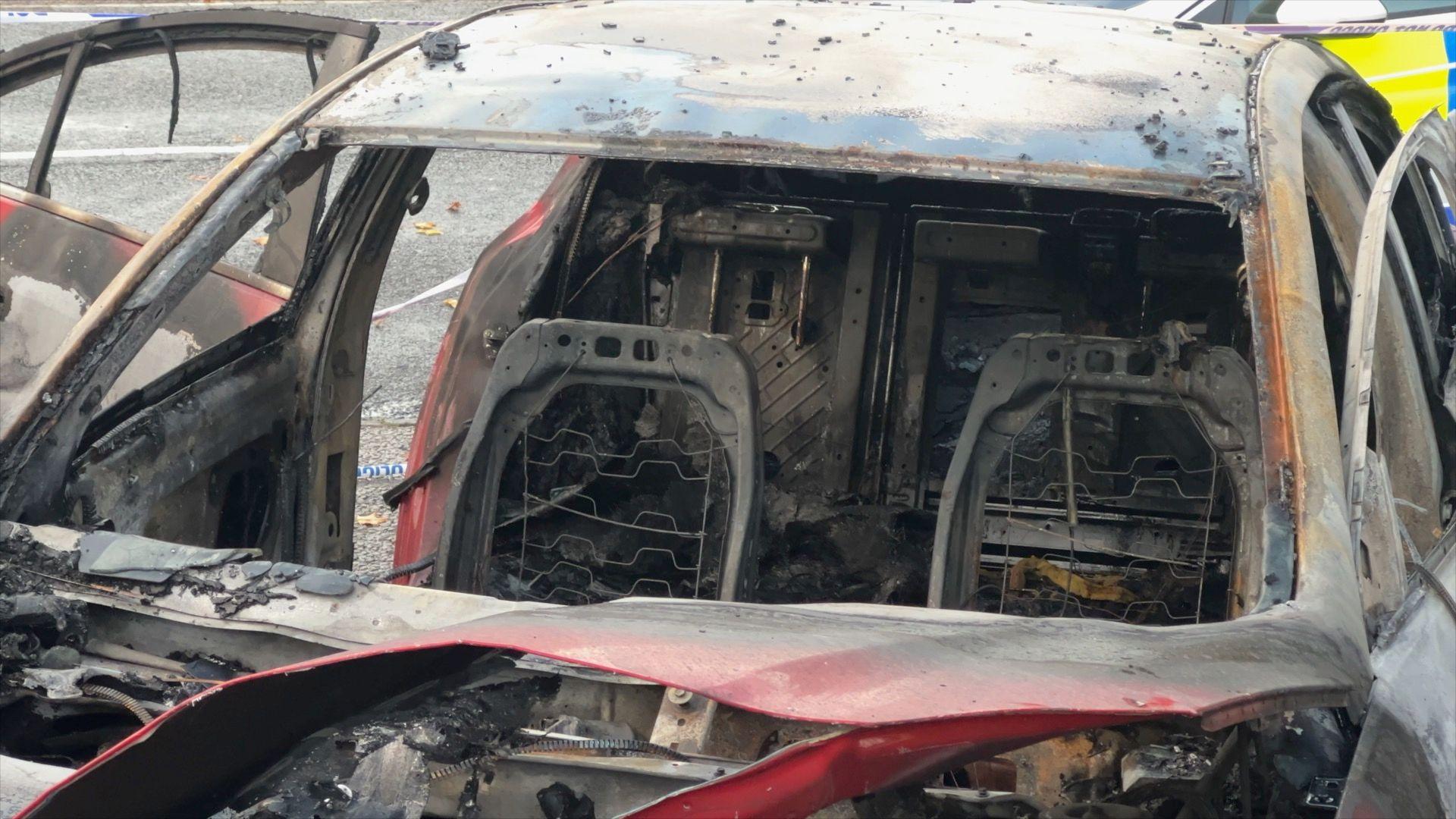 Front view of a red car that had been on fire. The front windscreen has been shattered and all that remains of the car seats is charred metal. The car is mostly charred black from where the fire has destroyed it. 