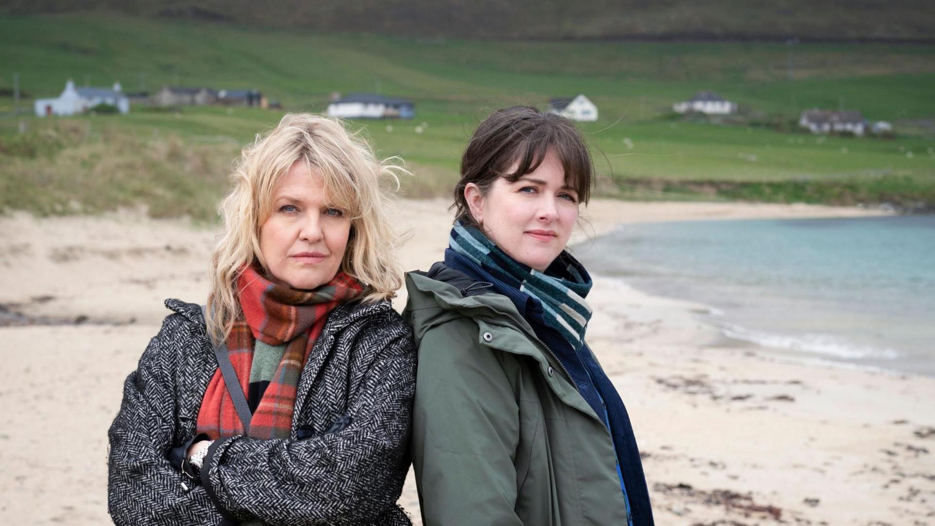 Actors Ashley Jensen and Alison O'Donnnell standing on a sandy beach. Both are dressed in warm, layered clothin. Ashley is wearing a textured grey coat and a red-and-green tartan scarf, and has her arms crossed. Alison is wearing a green coat with a dark scarf and has her hands in her pockets.
Further back there is a grassy area with scattered white houses and a dark hill rising in the distance.