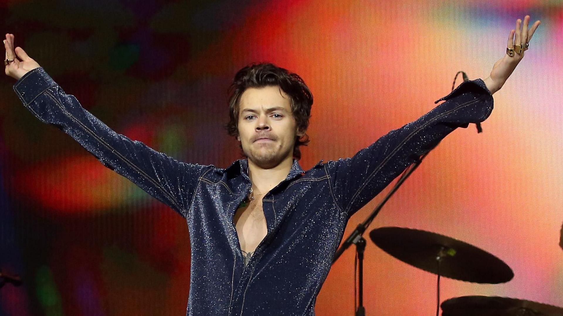 Harry Styles with his arms open wide. He is wearing a dark blue coloured top with white sparkles on it. He is standing in front of an orange background with a drum cymbal behind him.