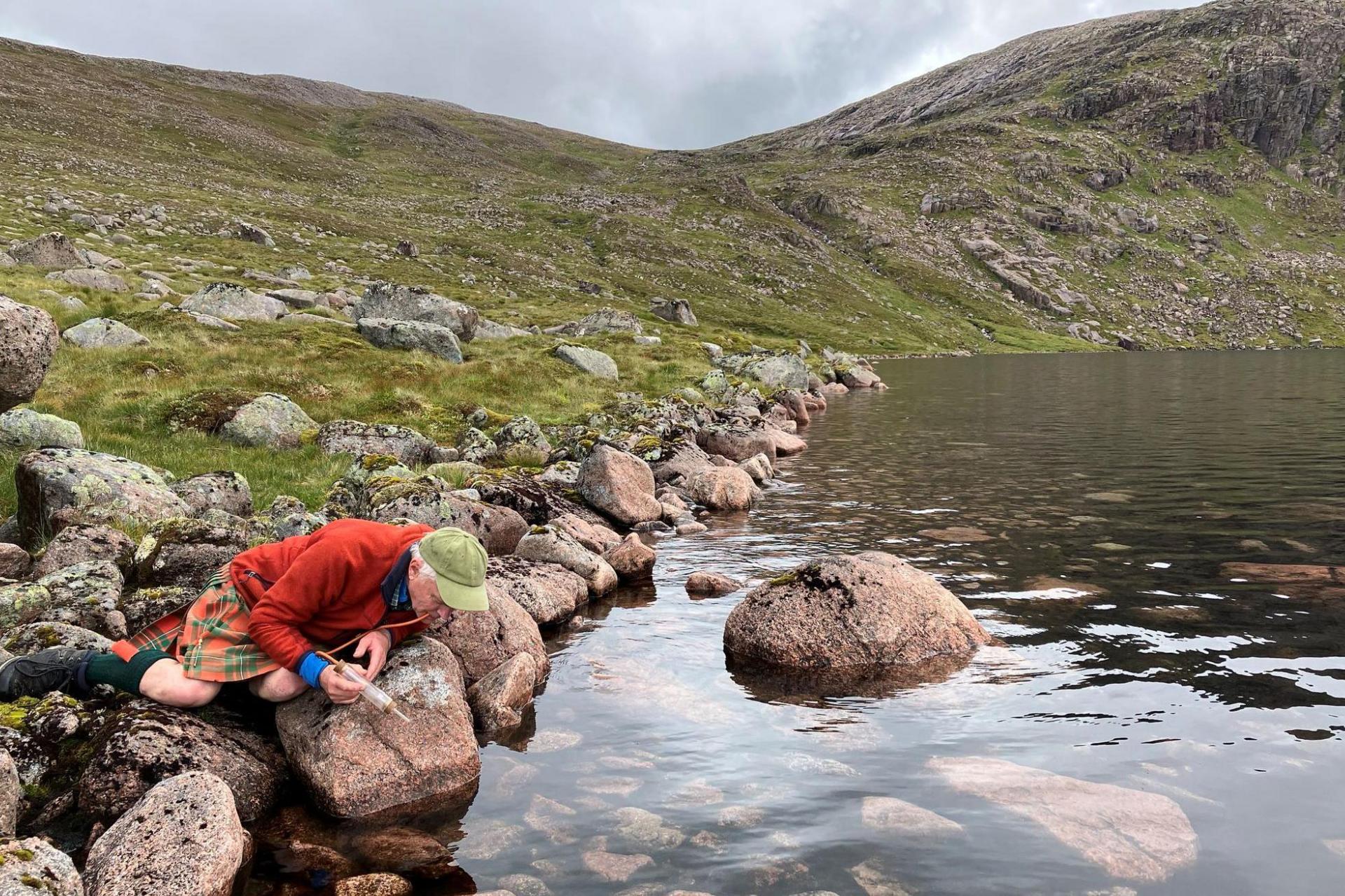 Iain, who is wearing a kilt and a green cap, is lying on rocks at the edge of the loch looking for arctic wave dancers.