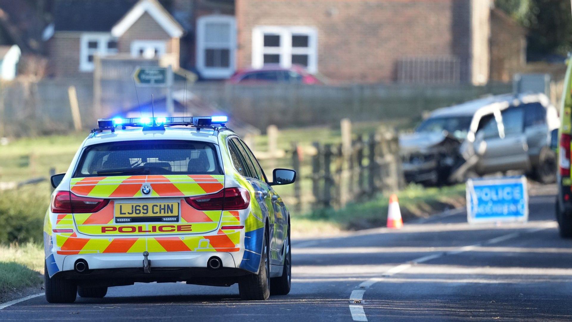 A police car is parked on a country road with a crashed car visible in the distance. 