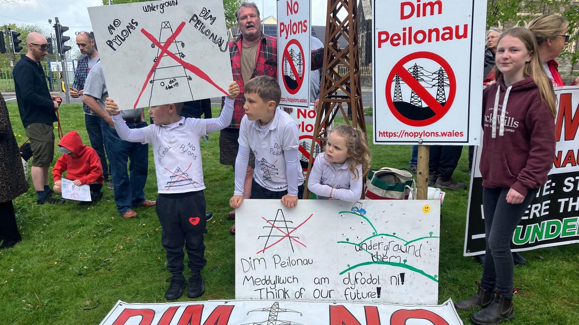 Protestors, including children, holding placards saying no pylons and dim peilonau, underground them standing on grass