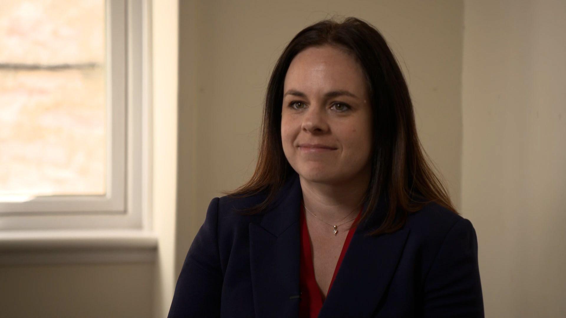 Kate Forbes - a young woman with long dark hair, seated and wearing a dark blazer and red blouse 