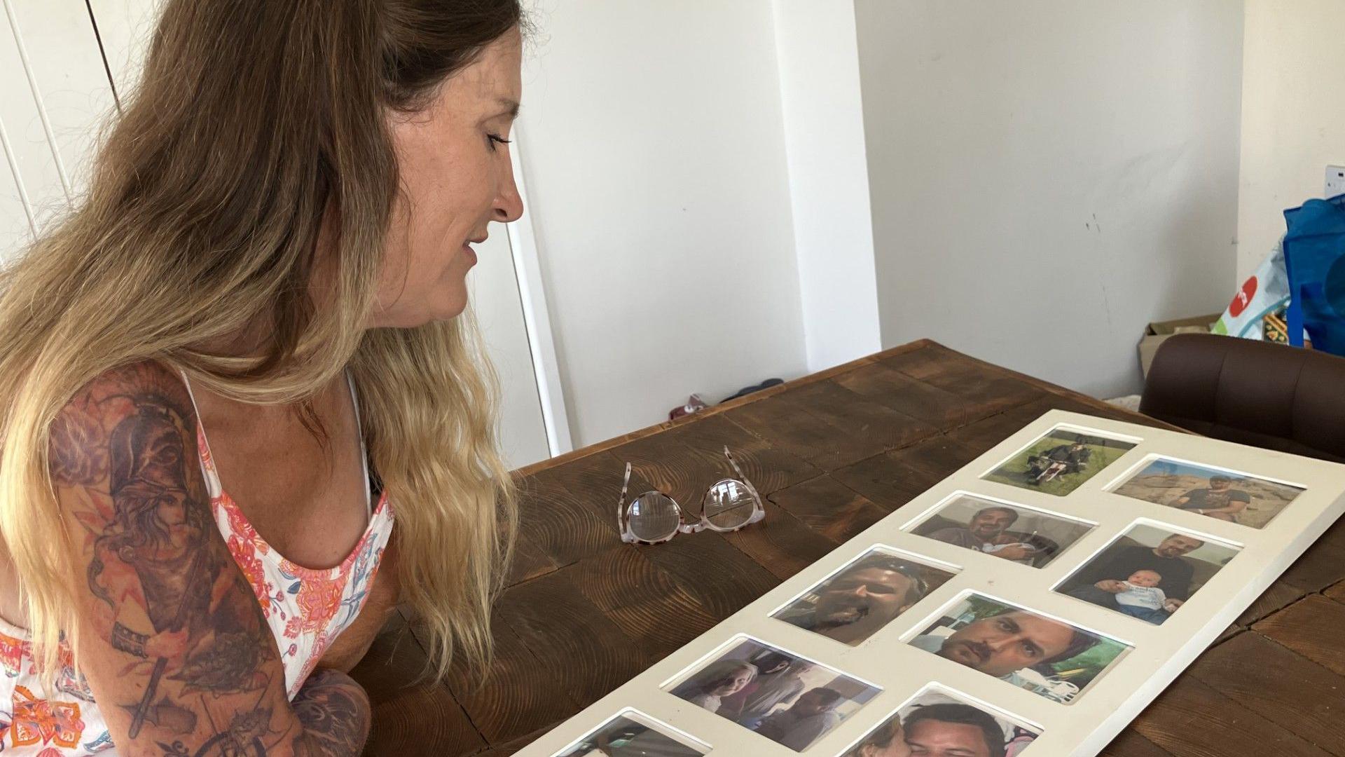 Caroline looks at a collage of photos featuring her husband Simon. The collage is resting on a table and Caroline is leaning on her arms looking down at it.
