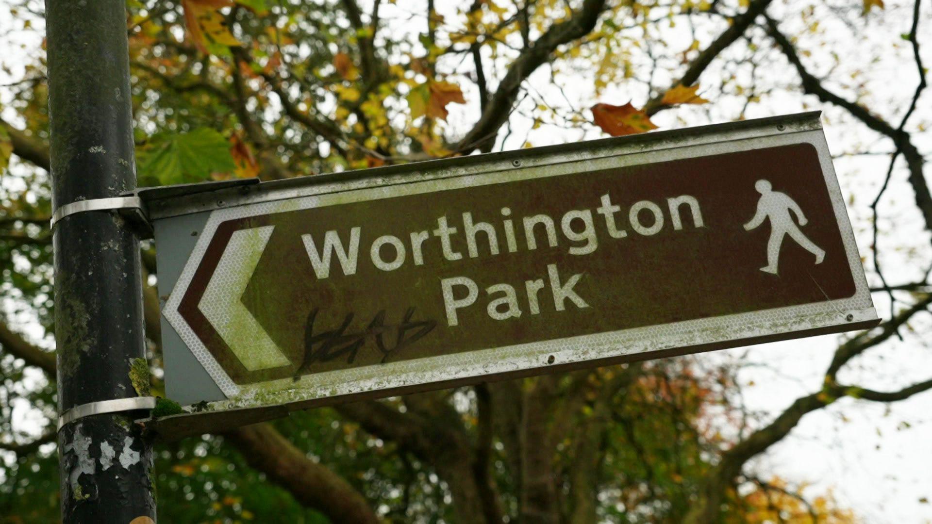 A brown arrow sign with white writing that reads Worthington Park. There is also a white silhouette figure of a person walking.