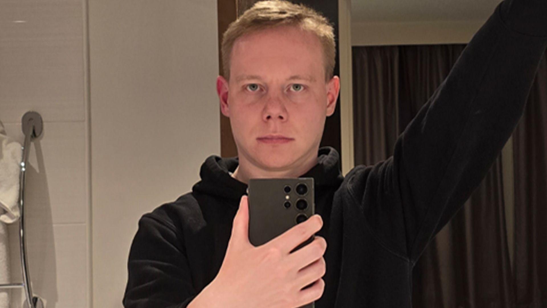 Parslow takes a mirror selfie in a hotel bathroom. He has short fair hair and is wearing a black hoodie.