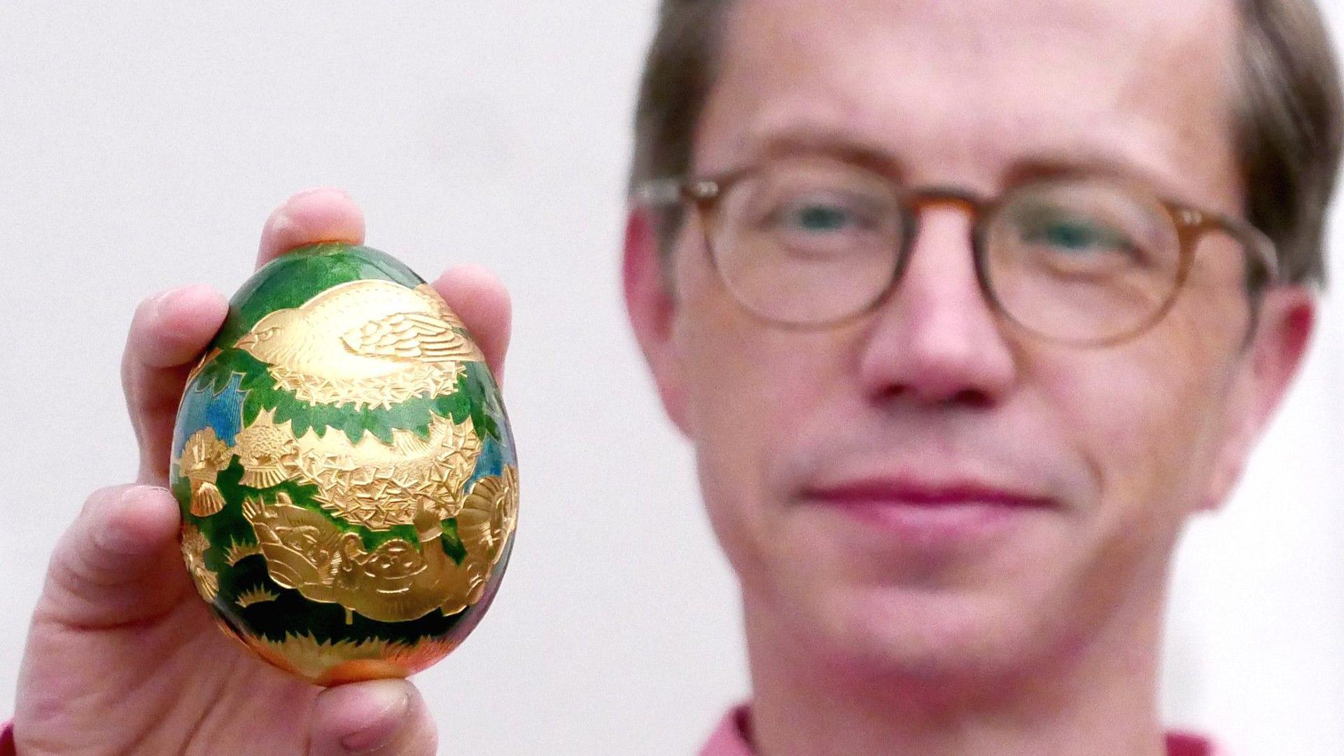 A green and gold decorative egg featuring a cuckoo design, being held up in one hand by a man wearing glasses out of focus in the background. 