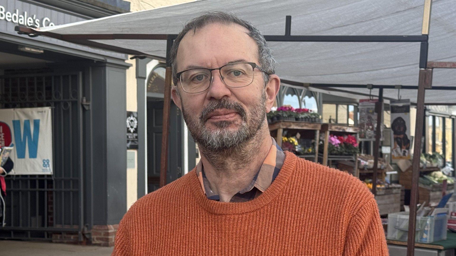 A man looks at the camera with a serious expression. He has greying hair, a moustache and a beard, and is wearing glasses. He's wearing a check shirt with an orange jumper over the top. A market stall is visible in the background.