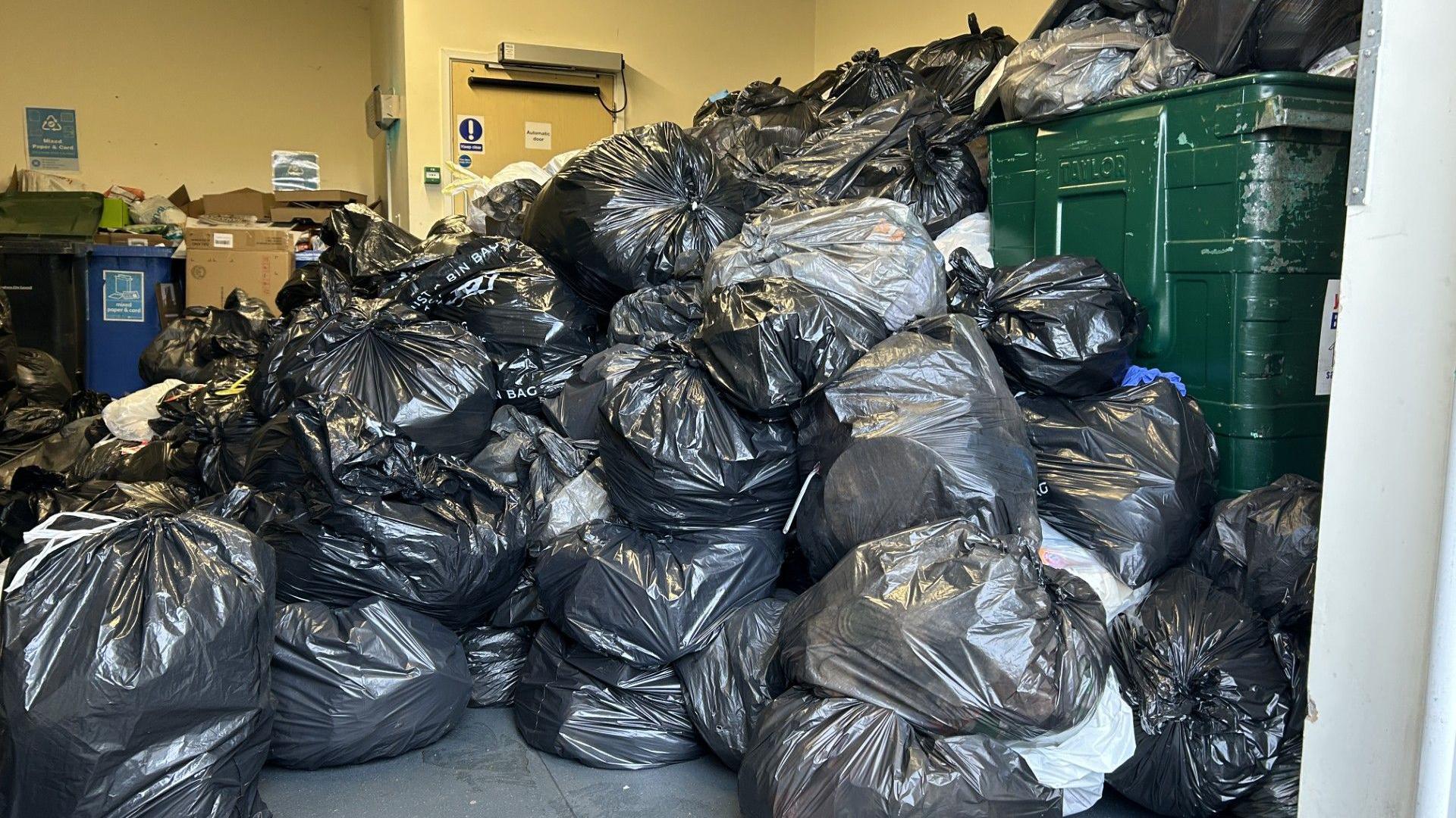 A huge mound of rubbish in a store room. There are lots of black and grey bins and containers full of rubbish bags behind them. Piled up recycling is away in the corner. 