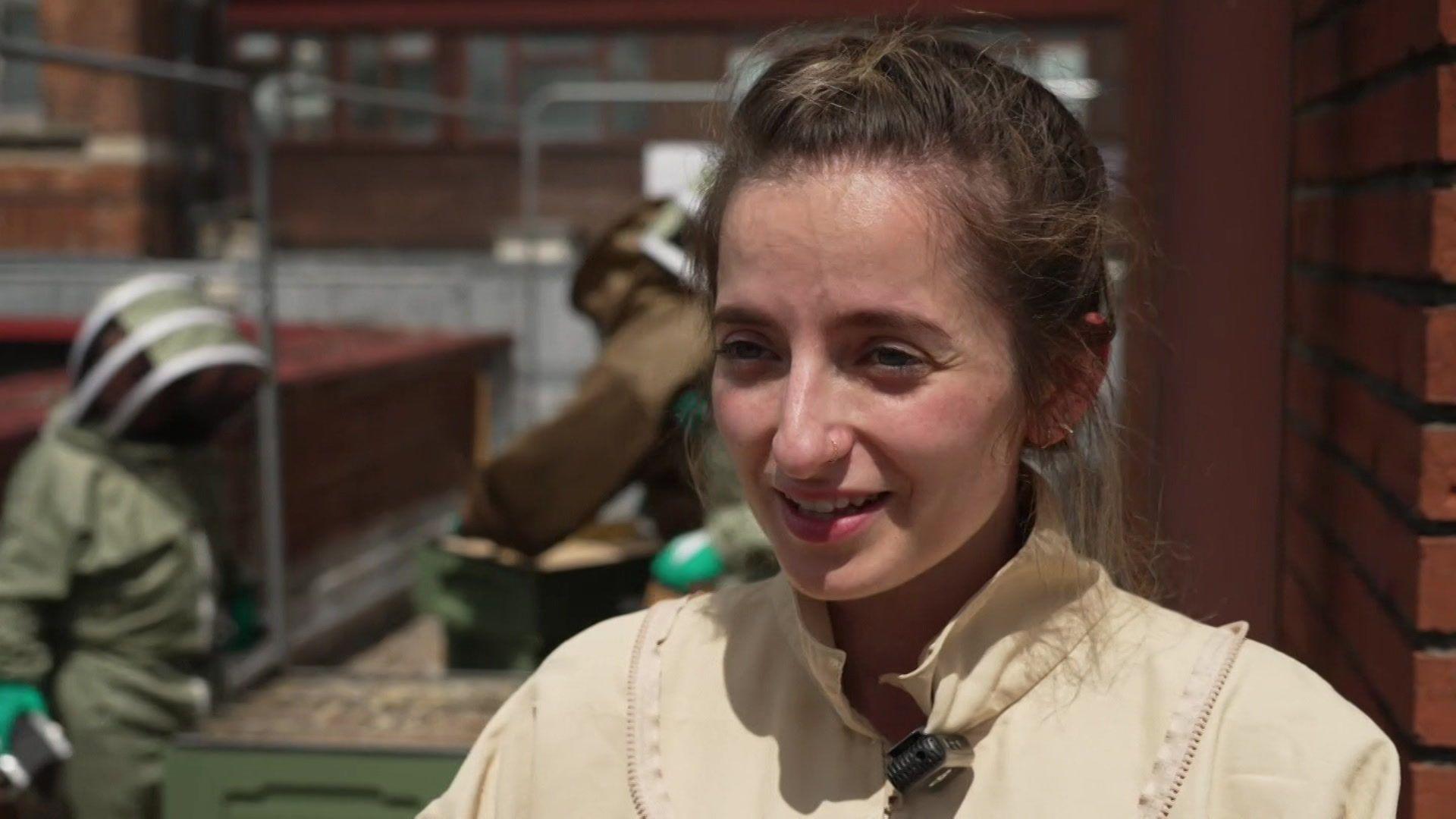 Christine wears a beekeeping suit. She has dark hair which is tied up.