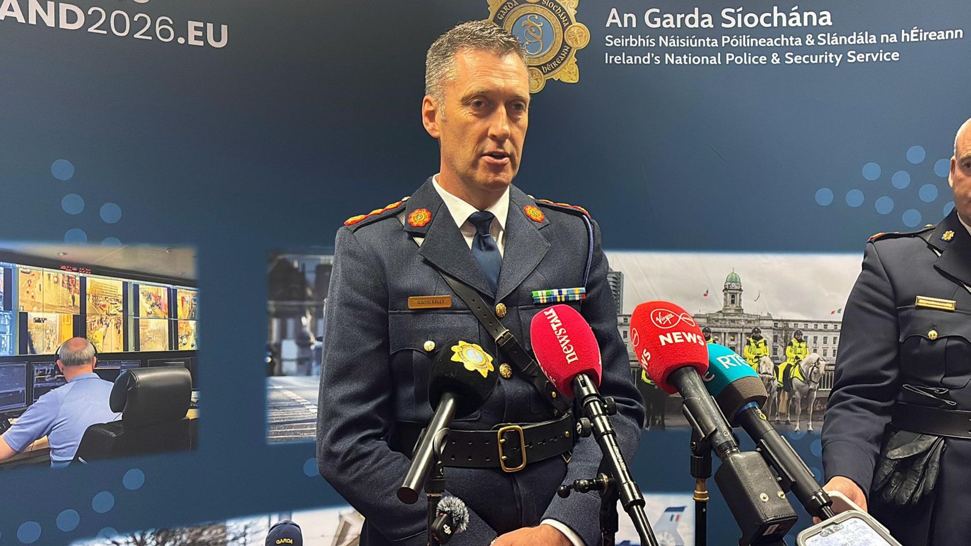 Garda officer wearing a blue uniform. He has short dark and grey hair and has mics in front of him. An Garda Siochana is on the background behind with police pictures.