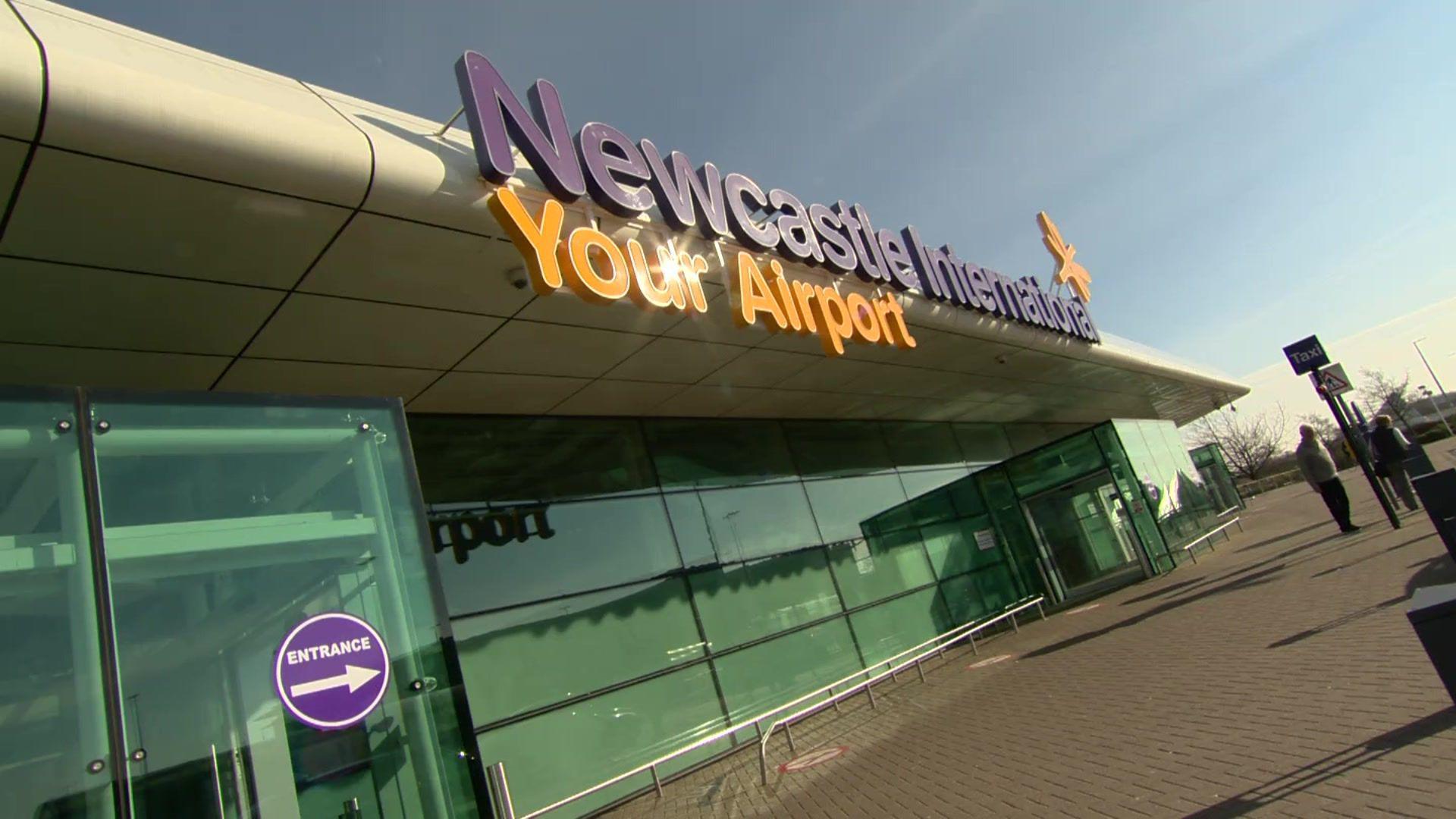 Newcastle Airport. A large glass building with a sign on the front of the roof reading 'Newcastle International Your Airport' in orange and purple lettering. There is a blue sky and two people in the distance standing on the pavement.