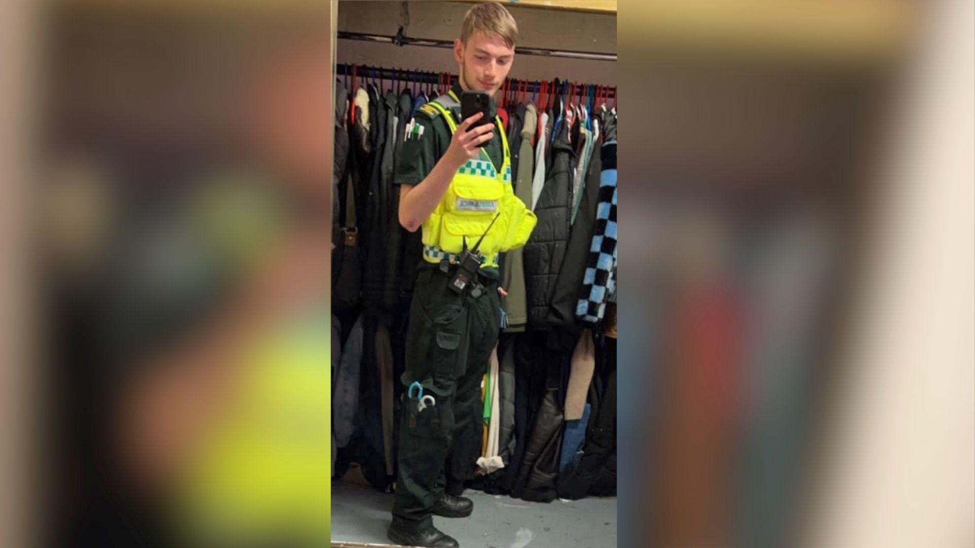 Jamie Kadolski poses for a selfie in a mirror. He is holding up a large phone with a black phone case. He is wearing a the uniform of a paramedic.