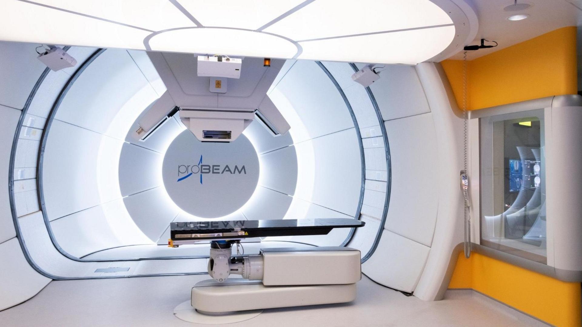 A futuristic-looking hospital room. There is a white circular backdrop, a shiny black surface to lie on, and instruments protruding from the ceiling above.