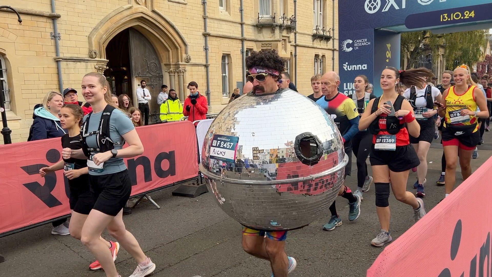 A group of runners with a man in the middle wearing an afro wig and stood inside of an enormous disco ball.