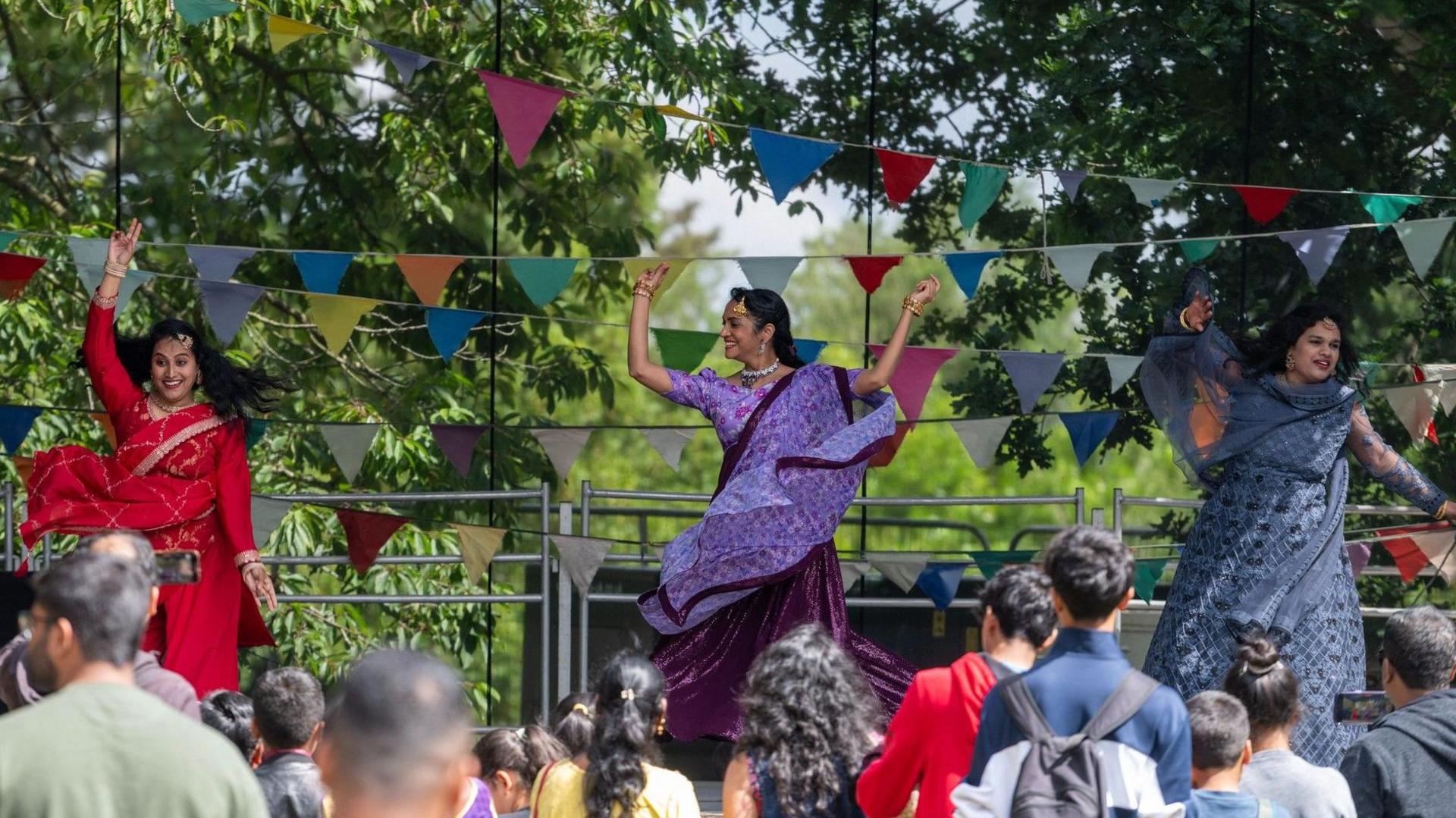 Two women in purple saree dresses dance on stage with their arms in the air. A crowd watches them. Bunting has been placed across the back of the stage.