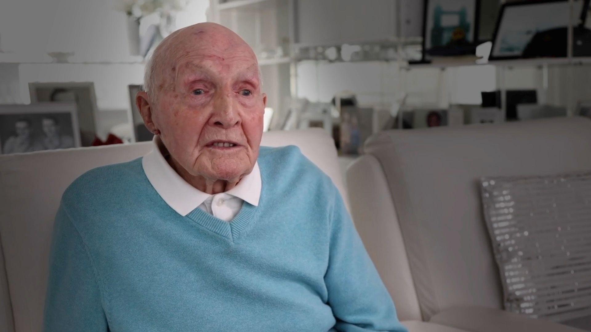 Leonard is bald, and is sitting on a white sofa. He is wearing a white shirt and blue jumper.