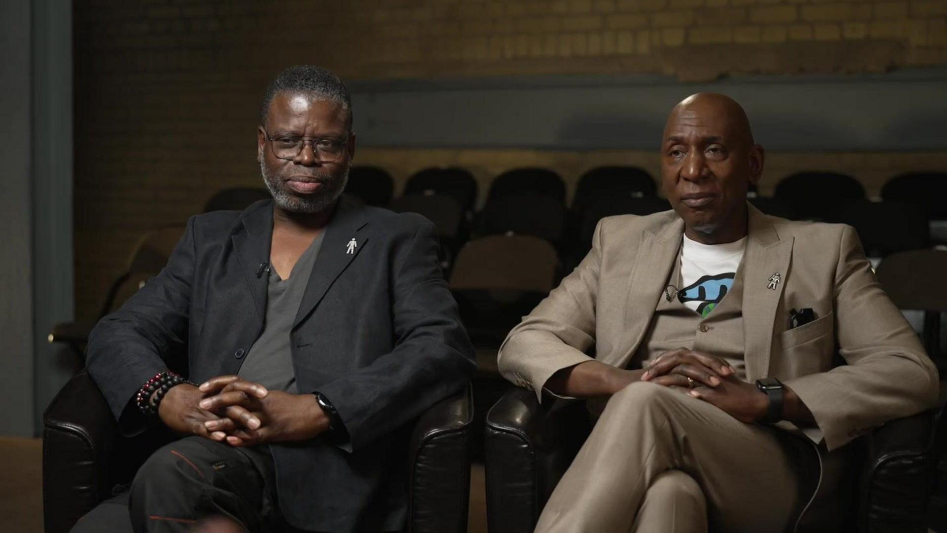 Femi and Colin are sat on two black leather chairs with their hands clasped in their laps. Femi is wearing a black suit and a dark grey t-shirt. He has rectangle glasses on and has short black hair. McFarlane is wearing a light brown suit with a light brown waistcoat and a white t-shirt with a blue, green and black pattern in the centre. He is bald. They are both sat in a dark theatre with rows of chairs behind them.