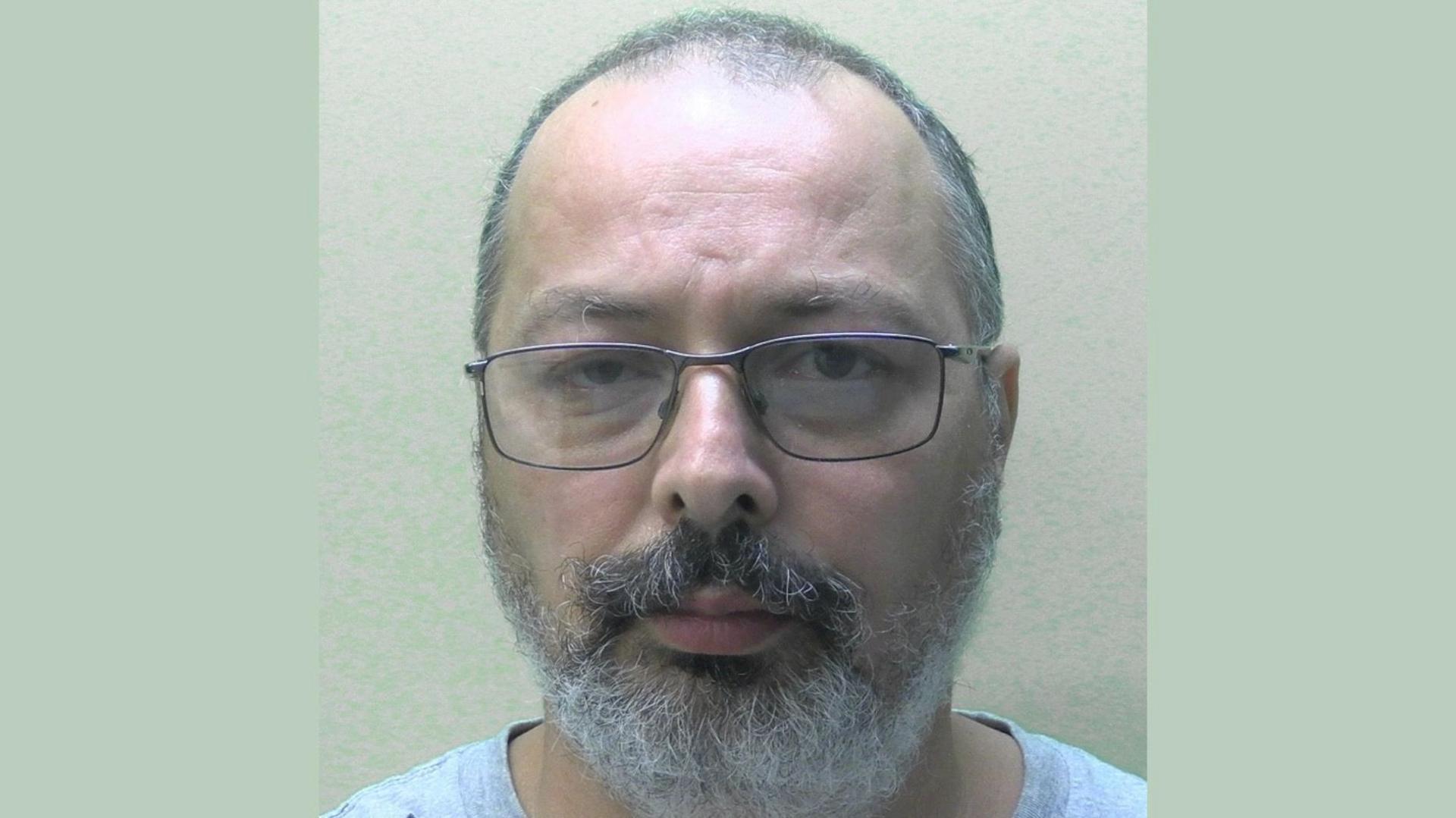 Richard Romeril wearing a tee shirt looking into the camera. He has a grey beard and moustache and is wearing metal-rimmed glasses