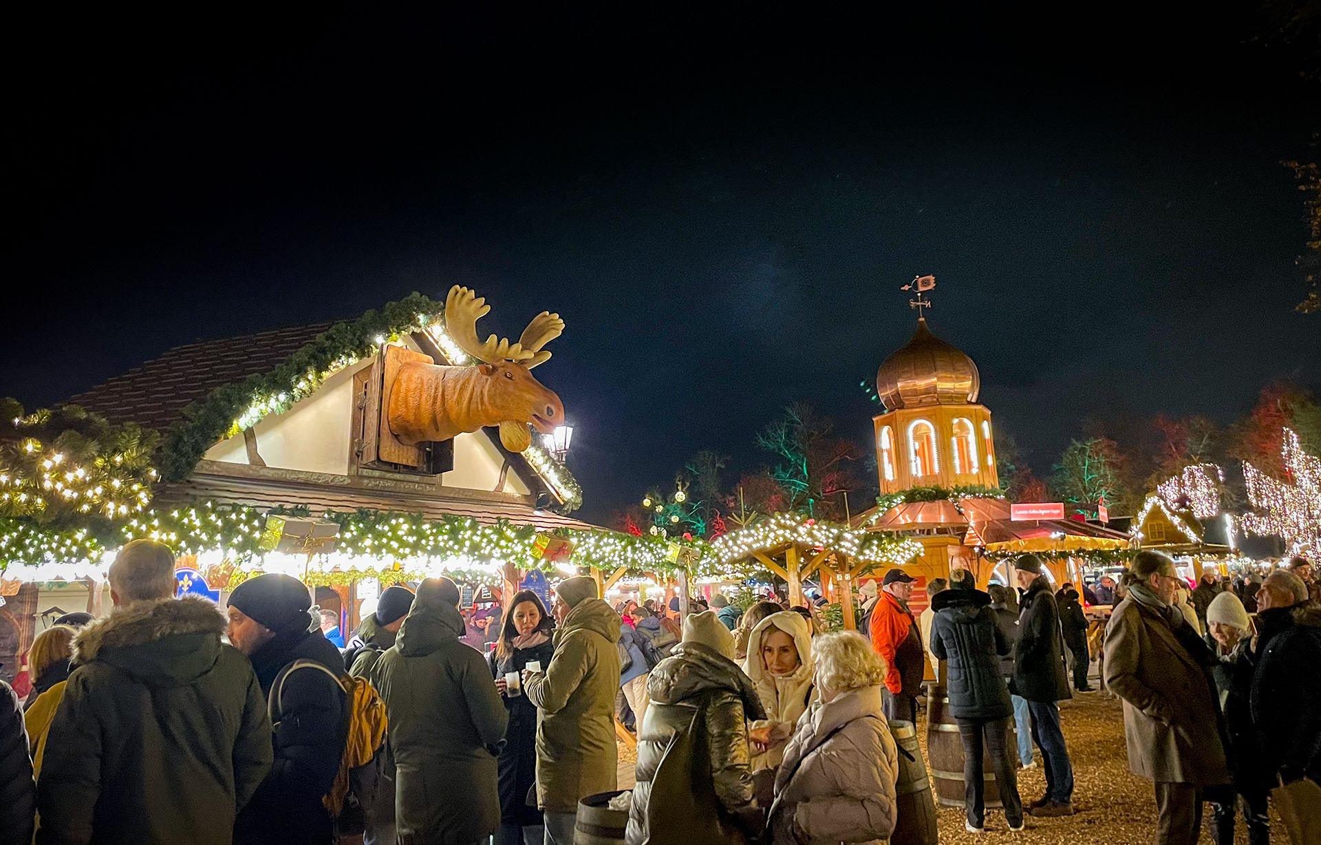 Crowds gather beneath treets lit up in red and green, lined with fairy lights, with wooden stalls in the background at a Christmas market in Berlin.