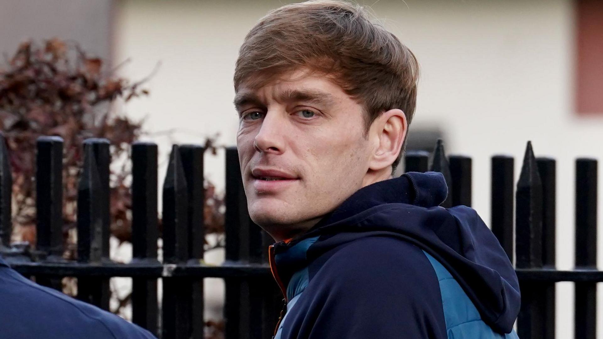 James Lawrence, who has light brown hair and wears a blue and navy coat. He has his back partially turned to the camera with his head tilted towards it.