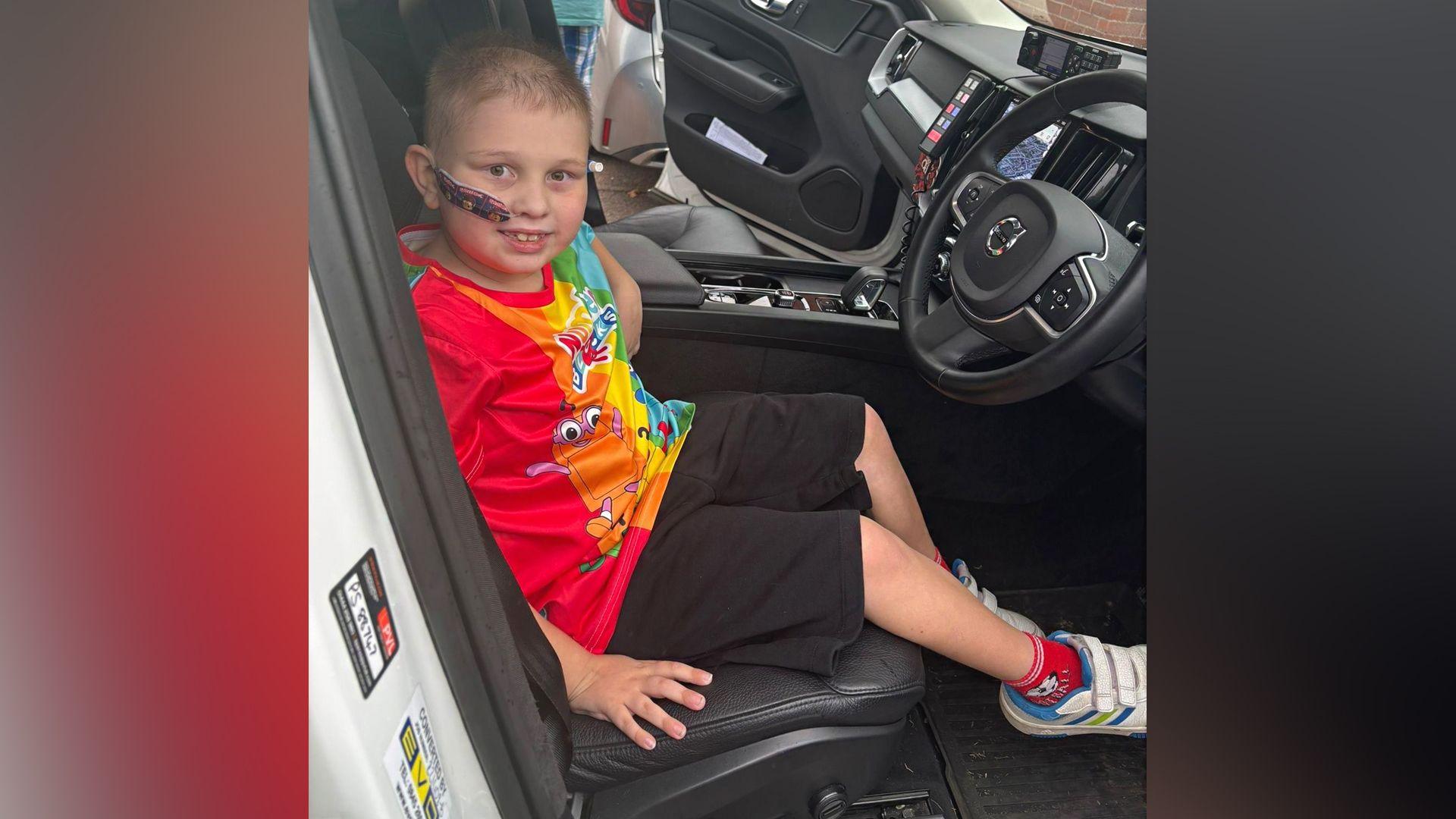 Luke is sitting in the front seat of the police car. He is wearing a rainbow coloured top, black shorts and white Velcro shoes. 