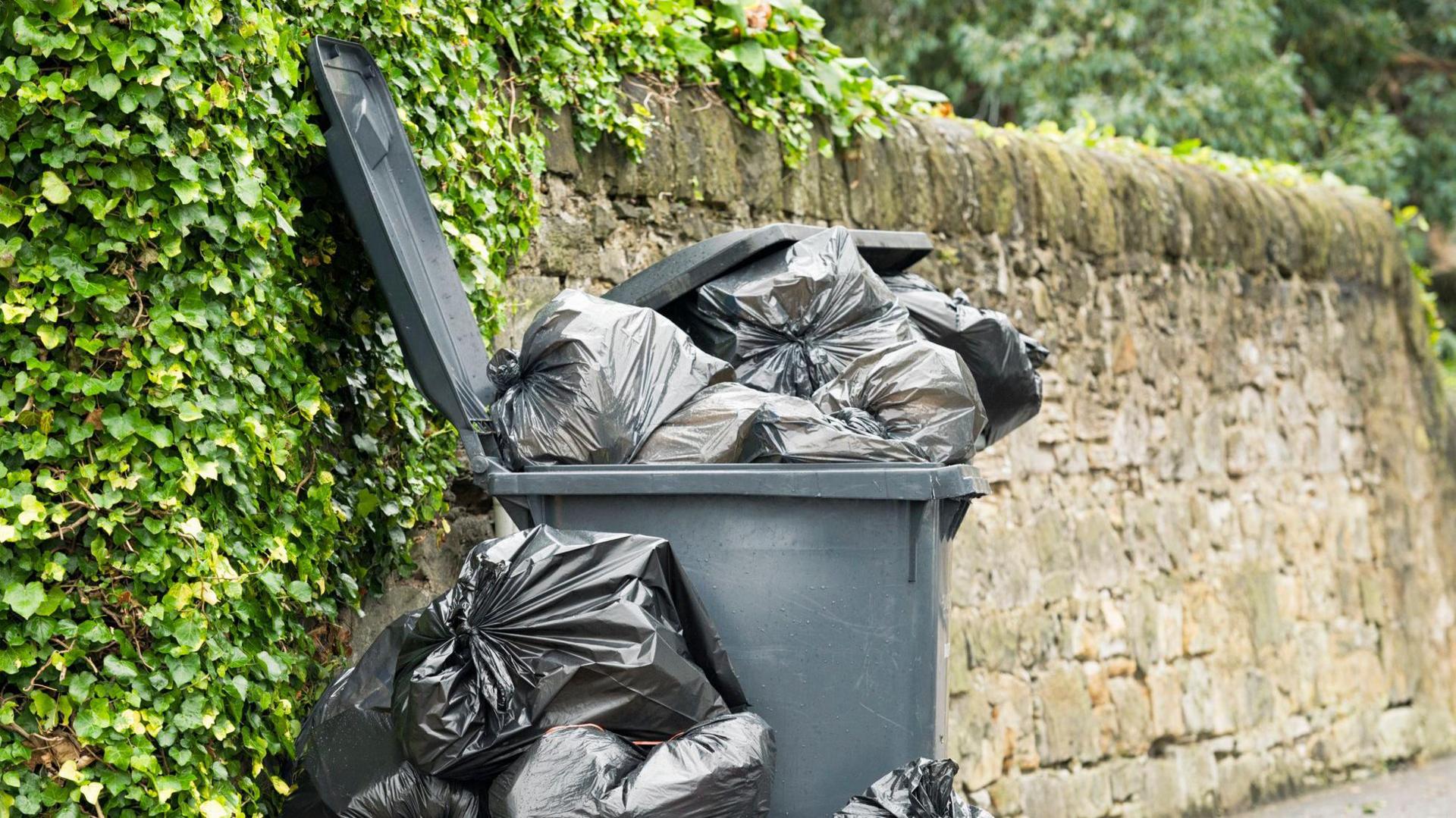 A black bin overflowing with plastic bin bags of rubbish. There is also a pile of full bin bags next to the bin. It is against a high stone wall, which has ivy covering part of it.