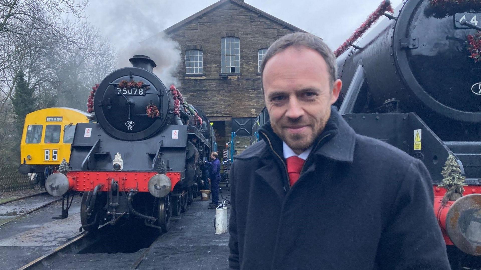 A man in a grey overcoat and wearing a red tie and white shirt underneath standing in the foreground with a large black locomotive with smoke coming out of it in the background.