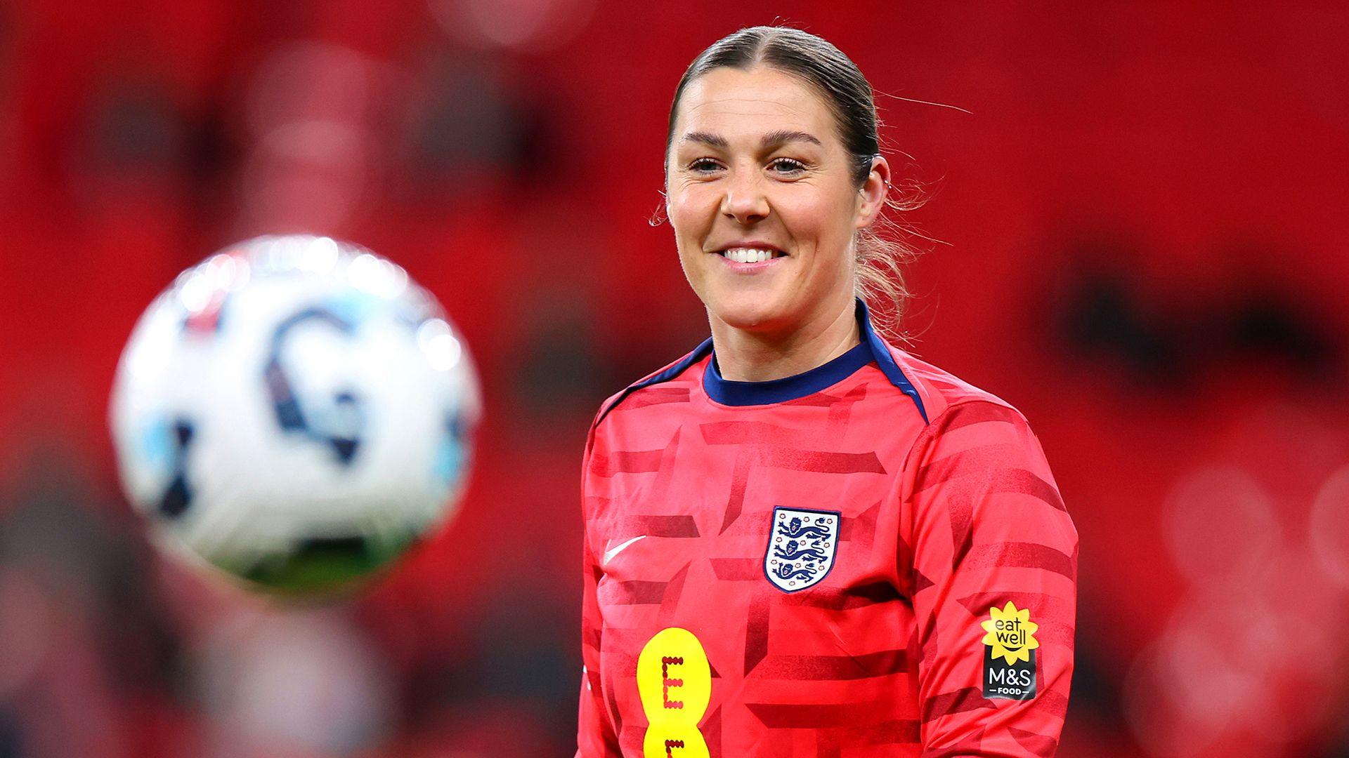 Mary Earps is pictured smiling with her hair tied back while a football moves in front of her in the foreground, she's wearing the red England football kit with out-of-focus red seats in the background, pictured at Wembley Stadium in February.