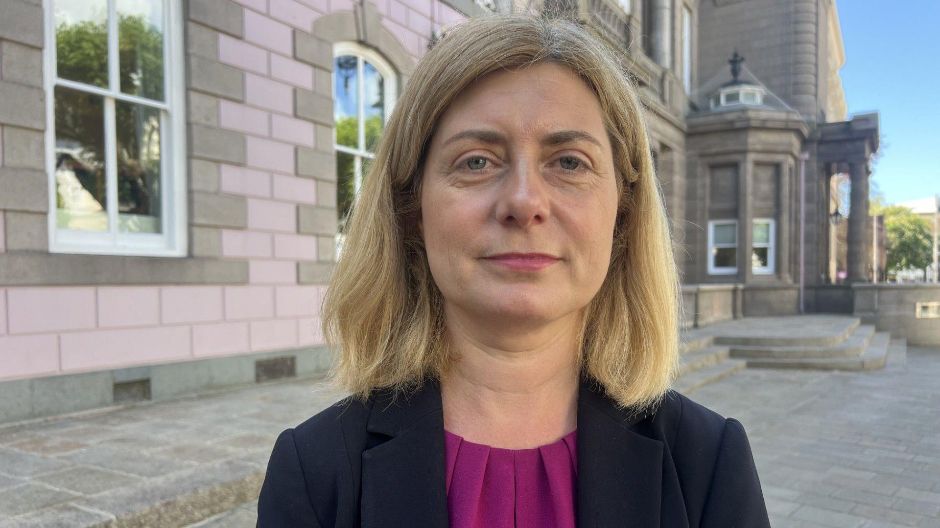 Deputy Lyndsay Feltham is standing outside a pink granite building on a  sunny day smiling at the camera. She has shoulder-length blonde hair and is wearing a navy jacket over a purple top.