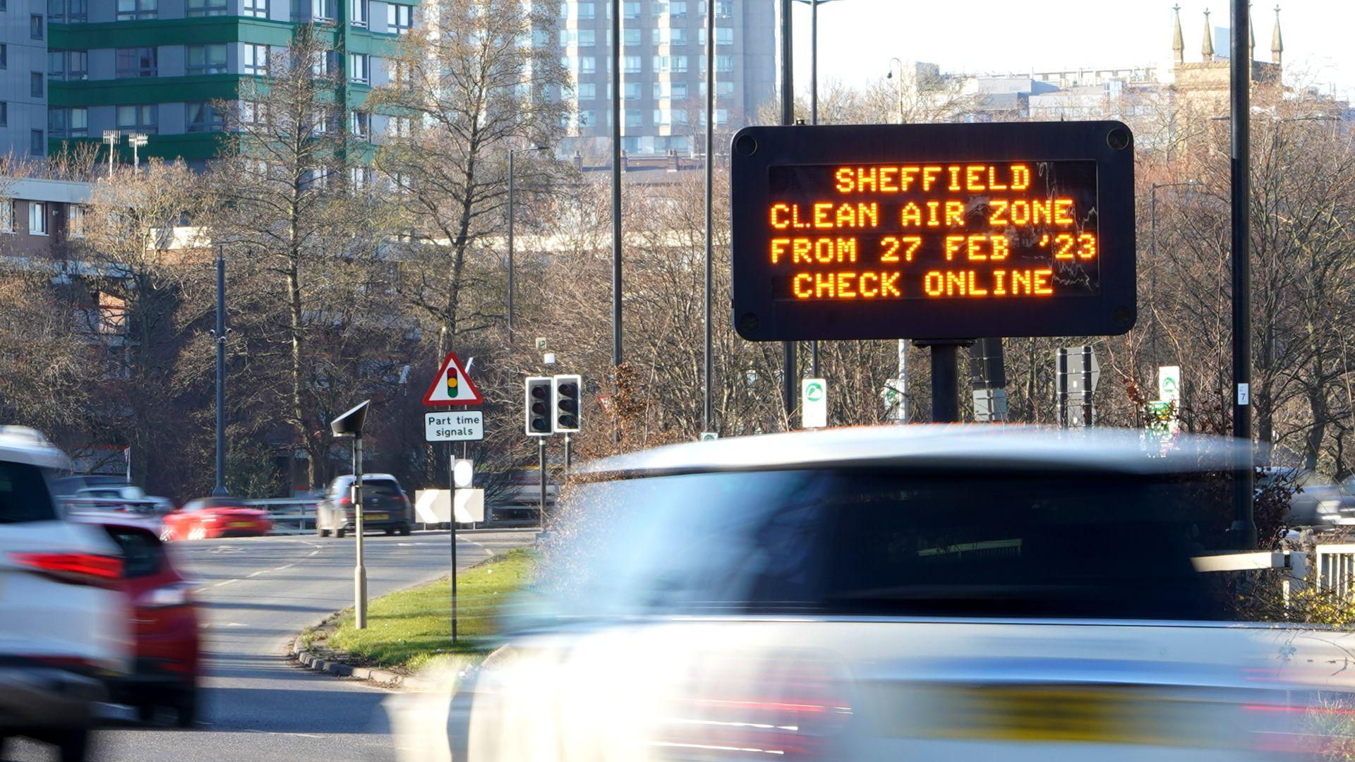 A busy road with a cream Mini blurred in the foreground as it travels along the road. An illuminated road sign reads Sheffield clean Air Zone from 27 Feb '23 Check Online. Trees and tall buildings in the background.