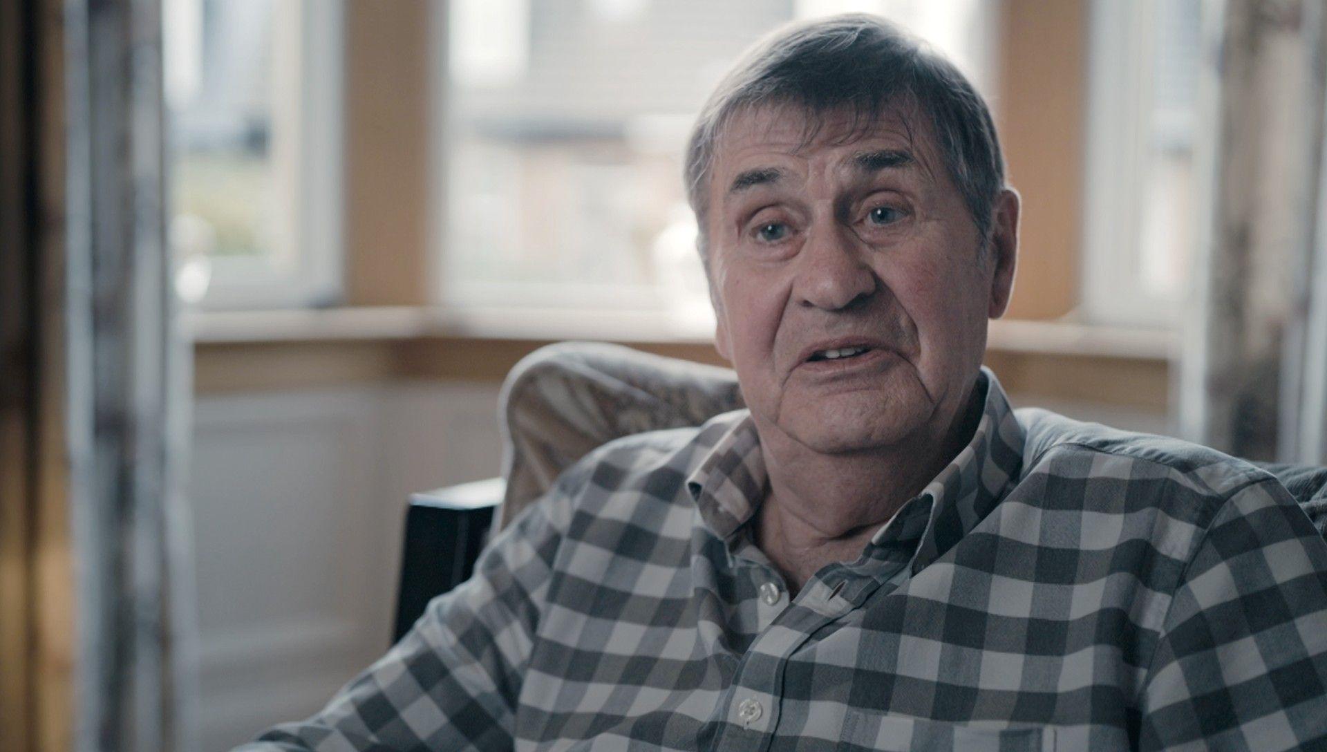 A man in his late 60s sitting in an arm chair in his home. He has dark/ grey hair and is wearing a checked shirt.