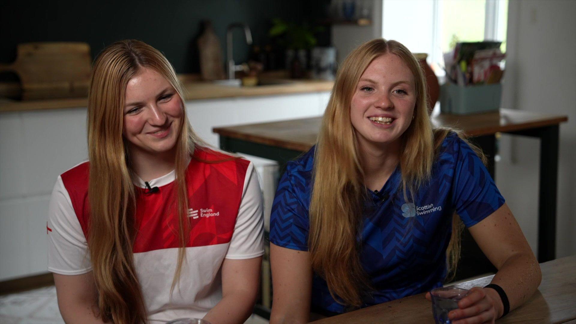 Astrid wears a blue Scotland top and Ursula sits next to her, wearing a red and white England top. They both have long blonde hair and are smiling. 