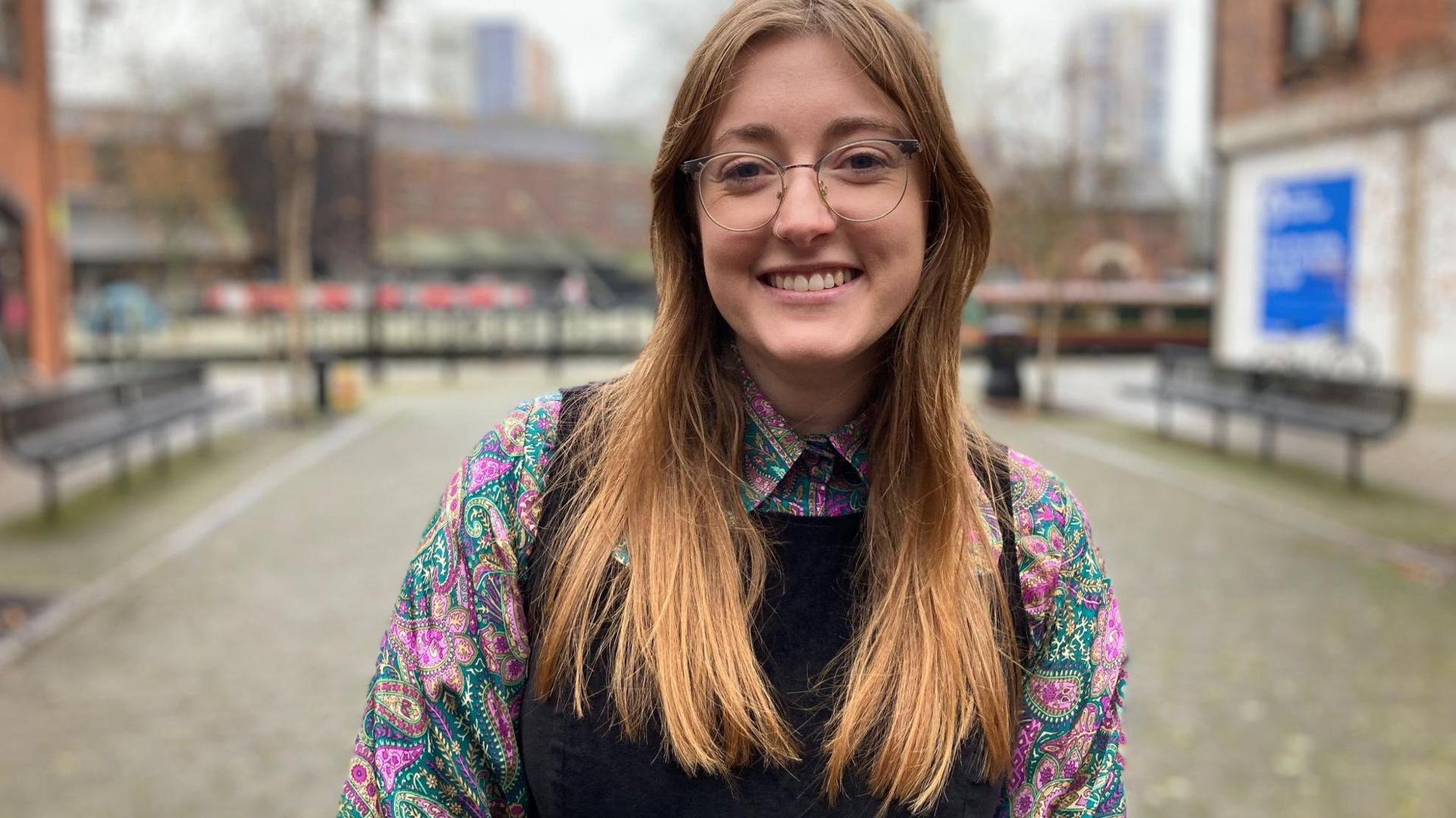 A woman smiling at the camera. She has long auburn hair and is wearing glasses and a patterned shirt and black dress. Behind her is a path and buildings which are out of focus. 