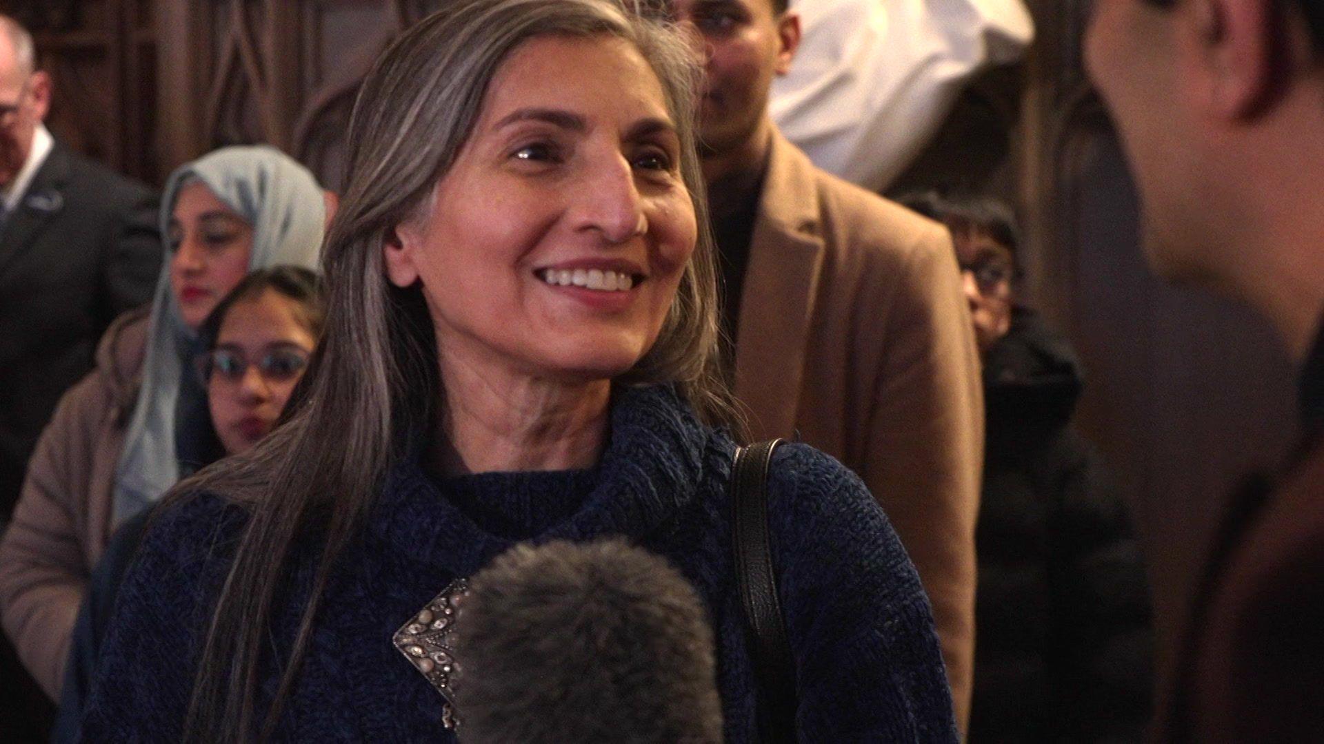 A woman in a navy blue jumper speaks to a journalist. behind her are other people attending the Iftar event.