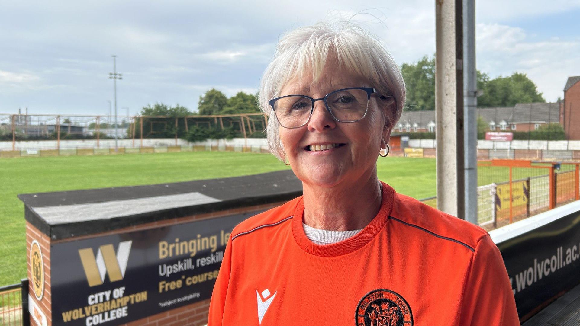 Kim Griffiths wearing a Bilston Town jersey. She is smiling at the camera. She has short hair and is wearing glasses. We can see the pitch behind her.